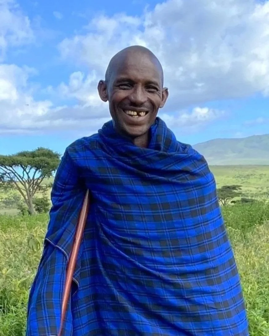 Smiling man with a gap-tooth smile in traditional Maasai attire, standing in a grassy landscape with trees and mountains under a partly cloudy sky.