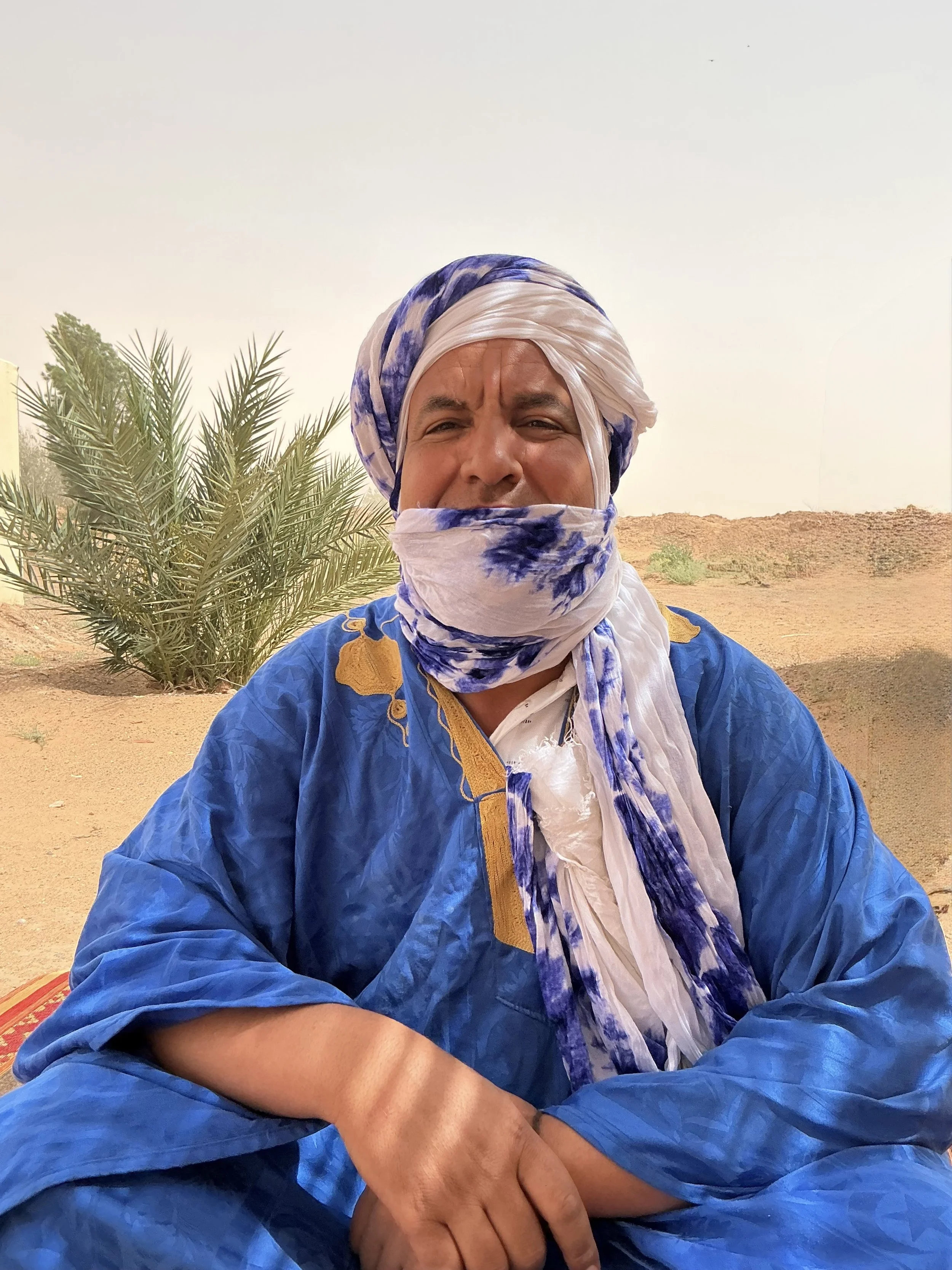 A man wearing a headscarf bends over a table with bags of supplies, bottles, and food items under a makeshift shelter in a desert environment.