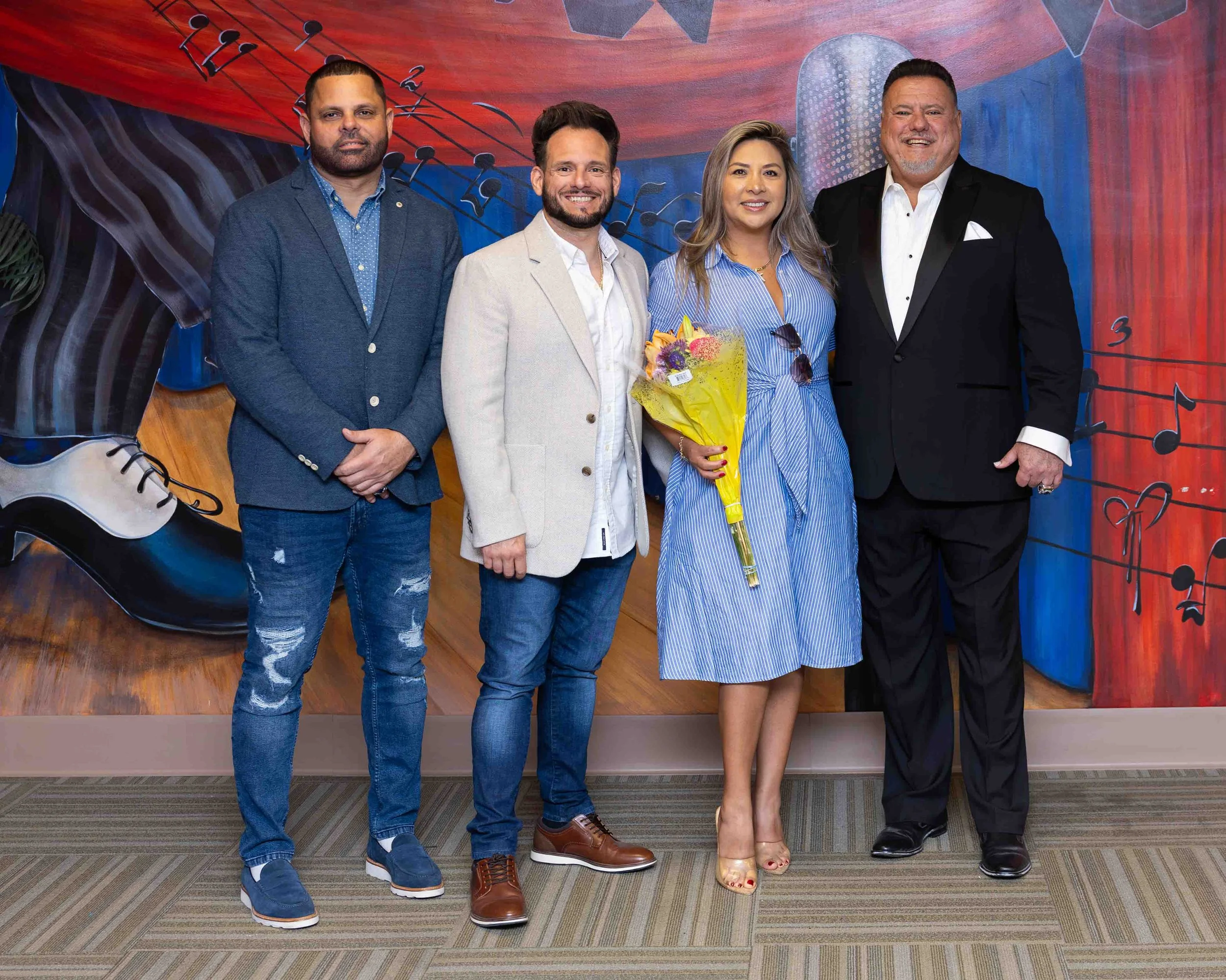 A group of five people standing in front of a colorful musical-themed mural, with one woman holding a bouquet of flowers, smiling for the photo.