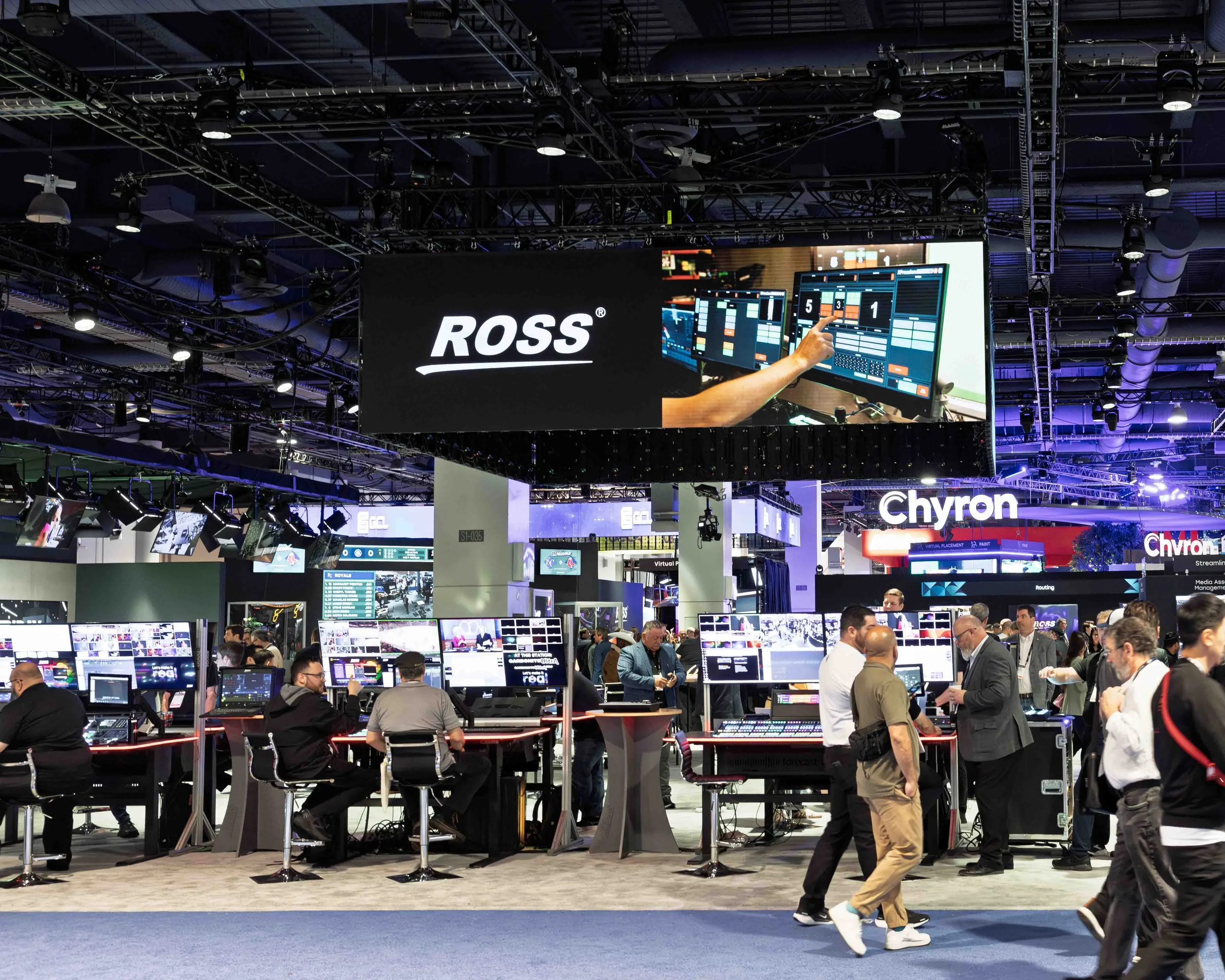A busy trade show or convention with numerous computer workstations and screens. Large branded signs for Ross and Chyron are visible overhead, and attendees are interacting and exploring various displays.