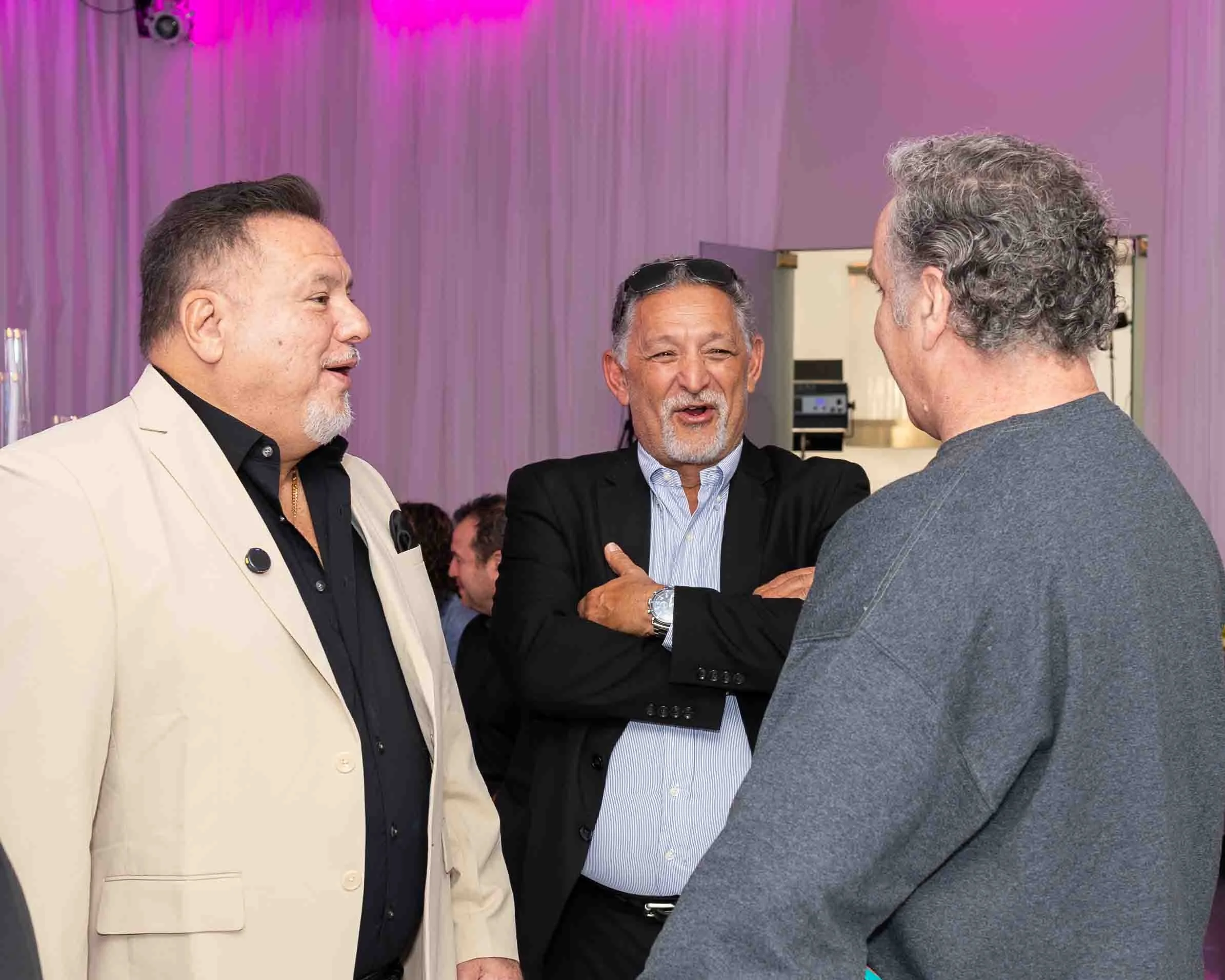 Three men are engaged in a conversation at a social gathering, with a pink-themed background and a partially visible kitchen area.