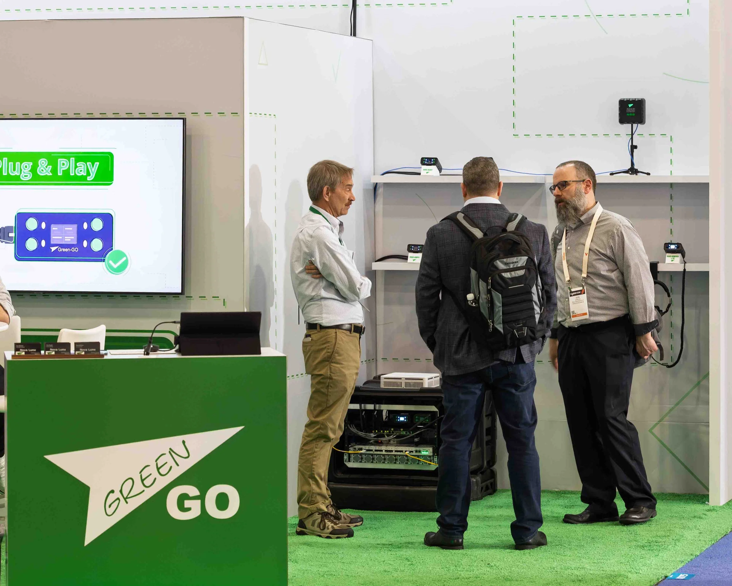 Three men having a conversation at a technology exhibit booth with electronic equipment on display. One man has a backpack, two are dressed in business attire, and one booth has a green and white sign reading 'GREEN GO' with a digital screen on the w