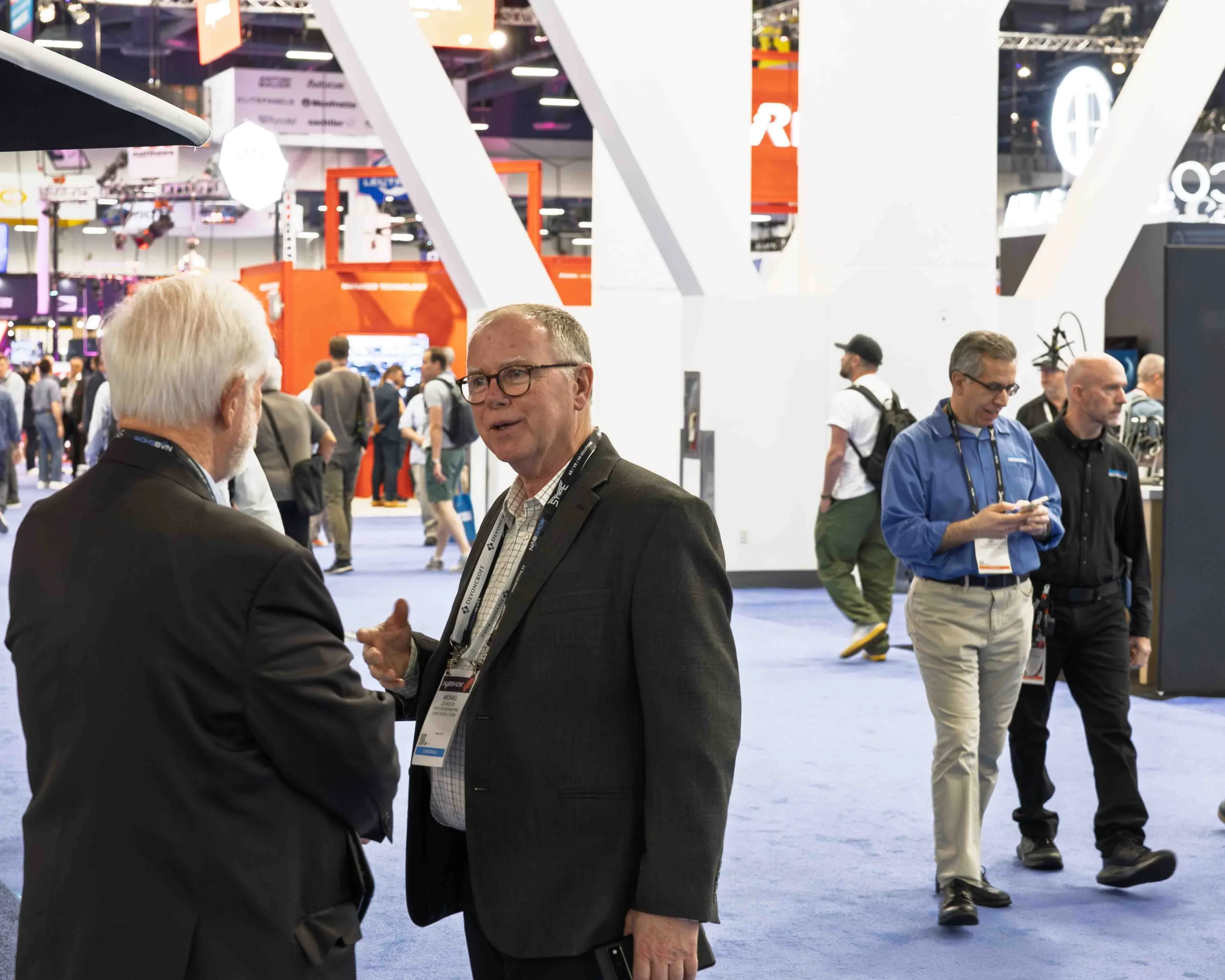 Two men are engaged in conversation at a busy trade show, with other attendees walking around in the background.