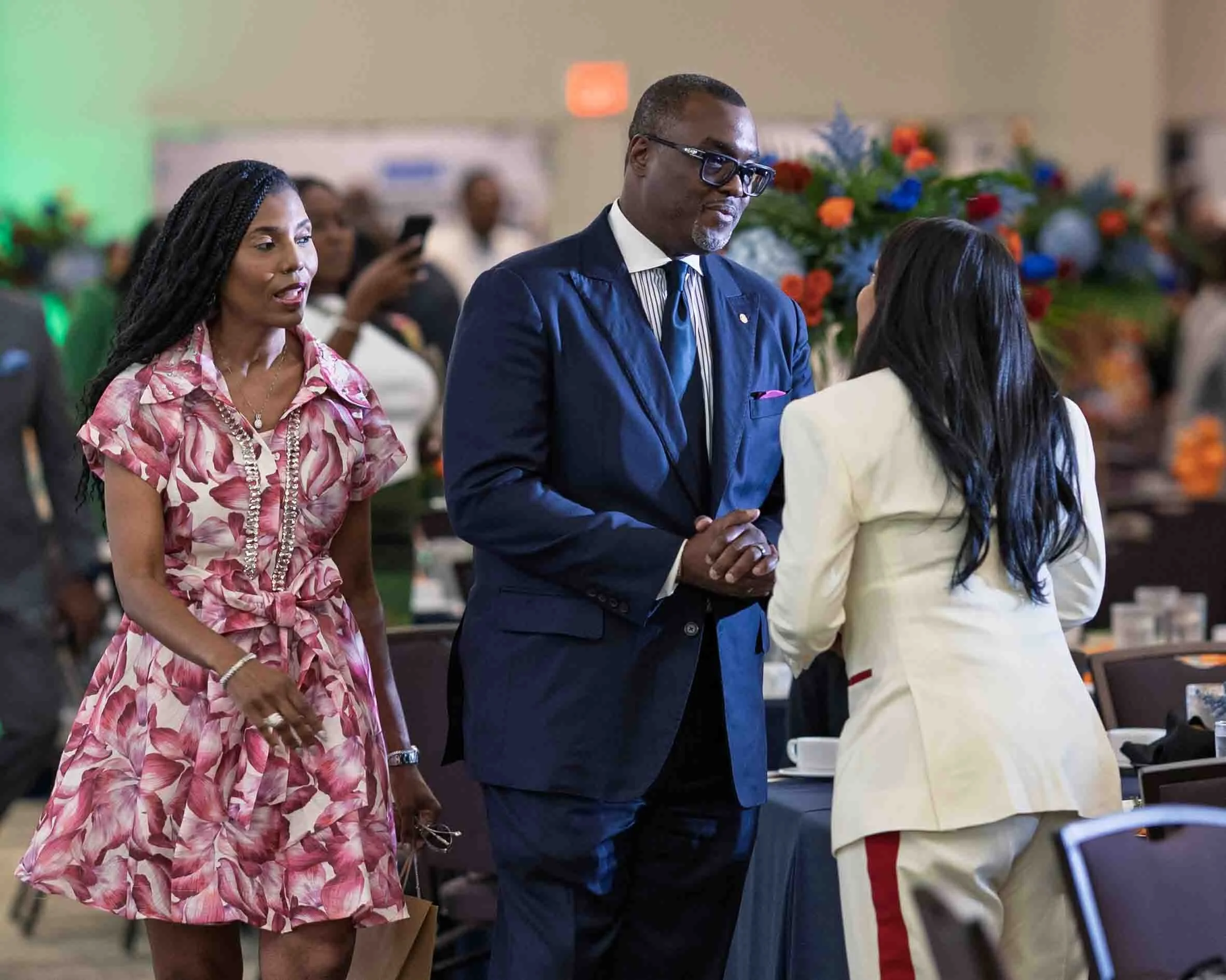 Three people engaged in conversation at a formal event, with colorful flowers in the background and other attendees in the distance.