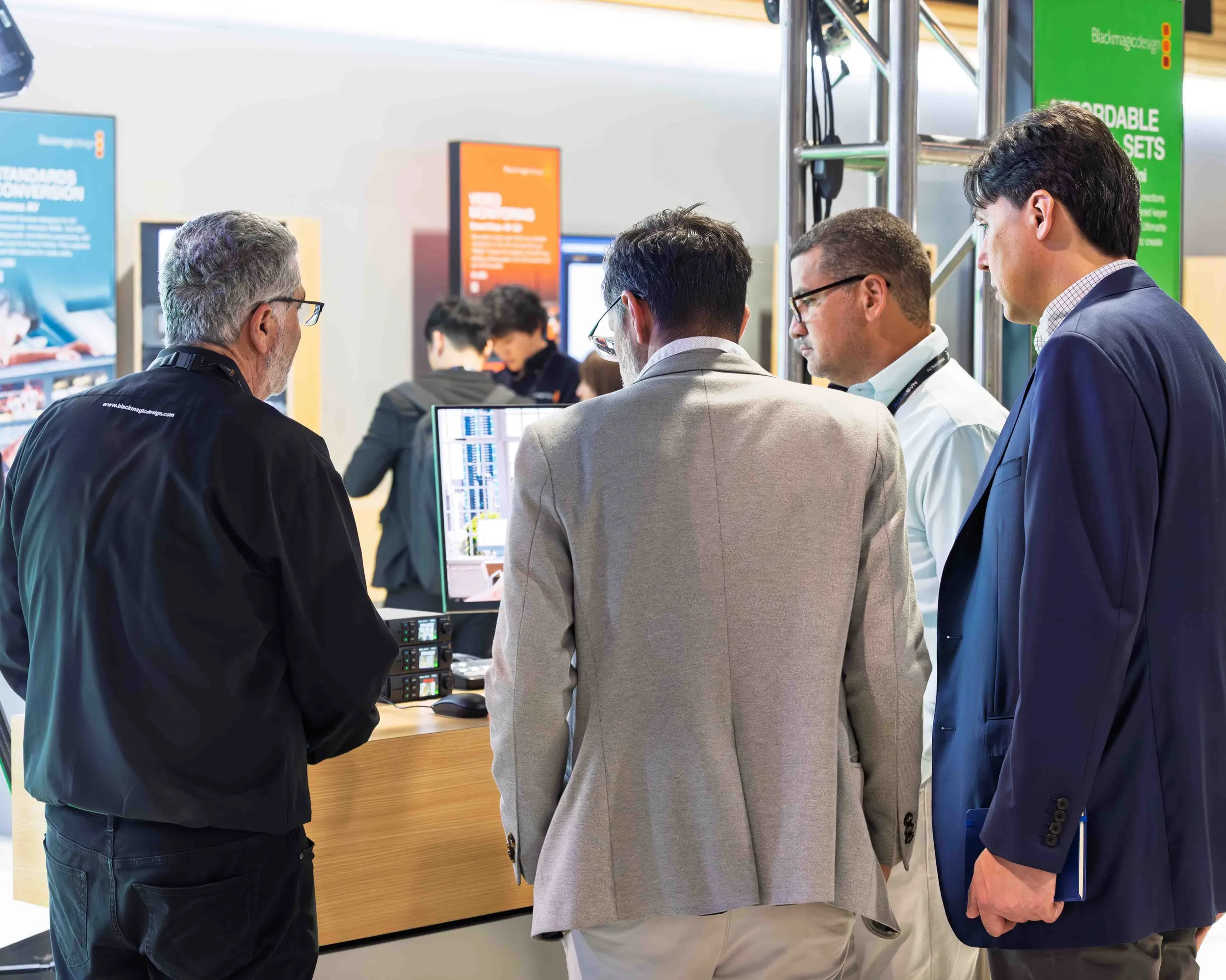 A group of five men in business attire gathered around a computer at a technology conference or exhibition. They are engaged in discussion, with some looking at the computer screen and others observing. Booth displays with colorful signage are visibl