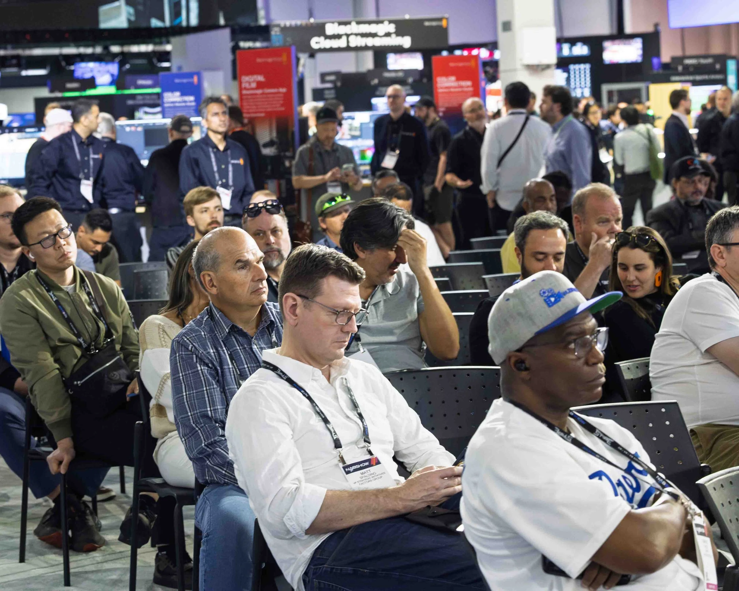 People attending a conference or seminar seated in rows with a busy exhibition or conference hall in the background.