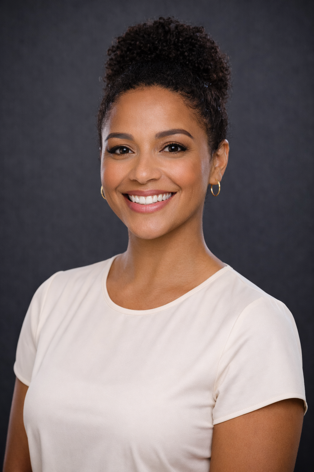 Headshot of smiling woman with curly updo hairstyle wearing a cream-colored top and small hoop earrings against dark gray background.