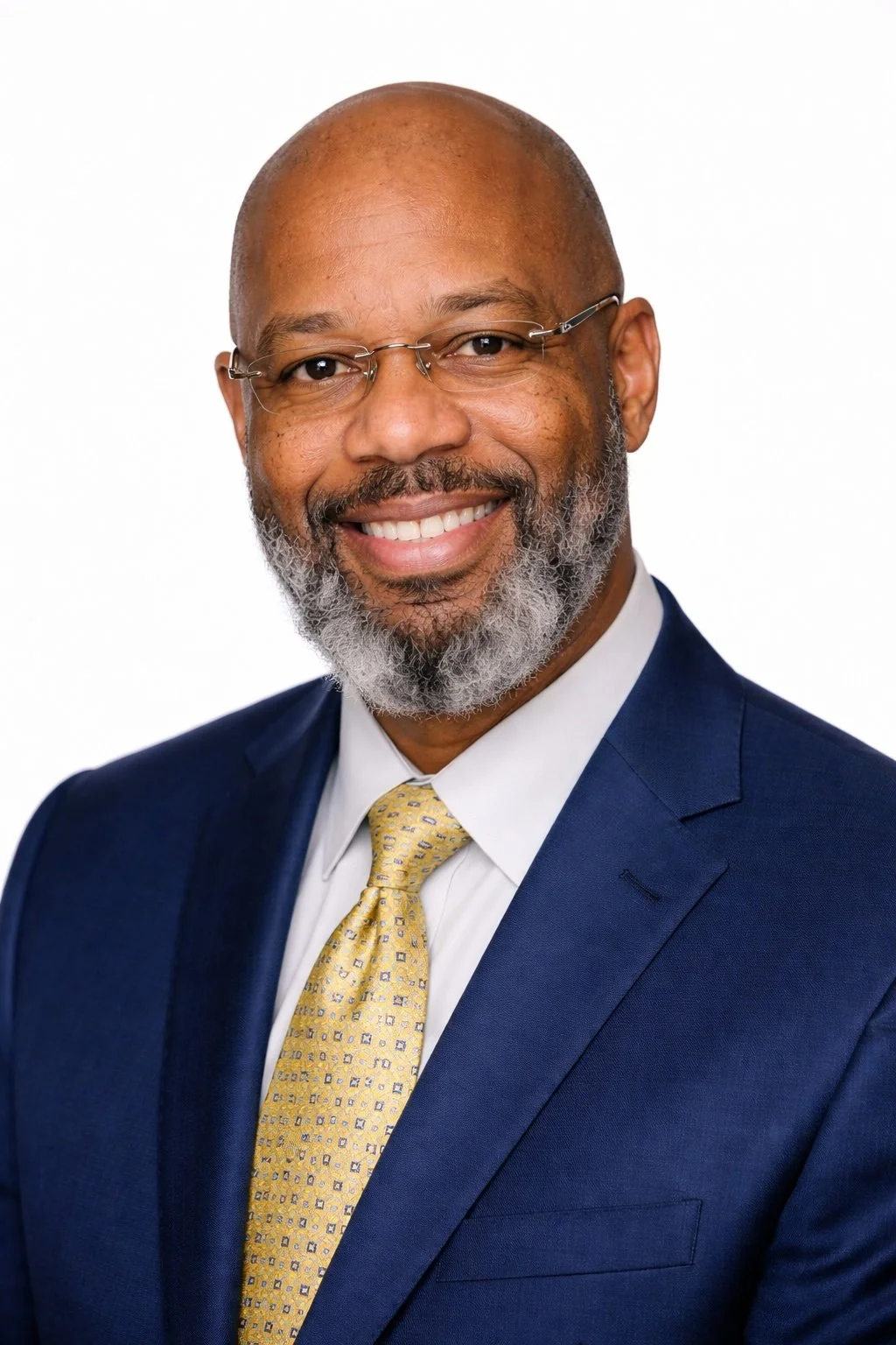 Headshot of a smiling African American man wearing a navy blue suit, white shirt, yellow patterned tie, and glasses against a white background.