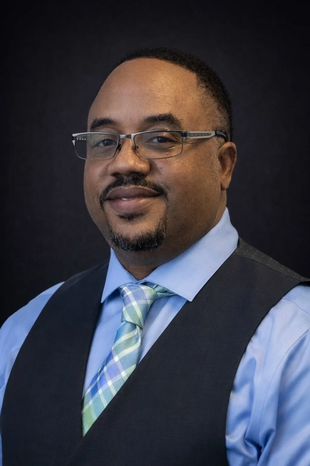 Headshot of a man wearing glasses, a light blue shirt, a patterned tie in pastel colors, and a dark vest, smiling against a black background.