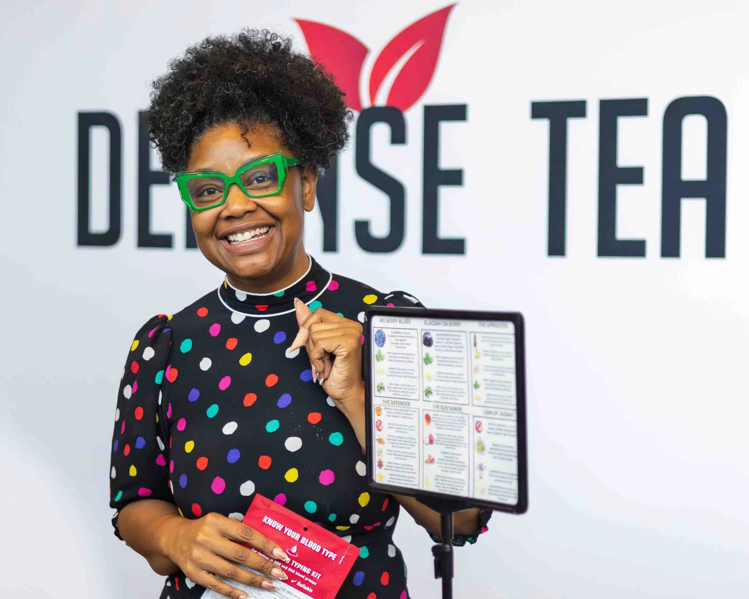 A smiling woman wearing green glasses and a black dress with colorful polka dots, holding a blood type reference card, standing in front of a white wall with the text 'DEFENSE TEA' and a red leaf logo at an event