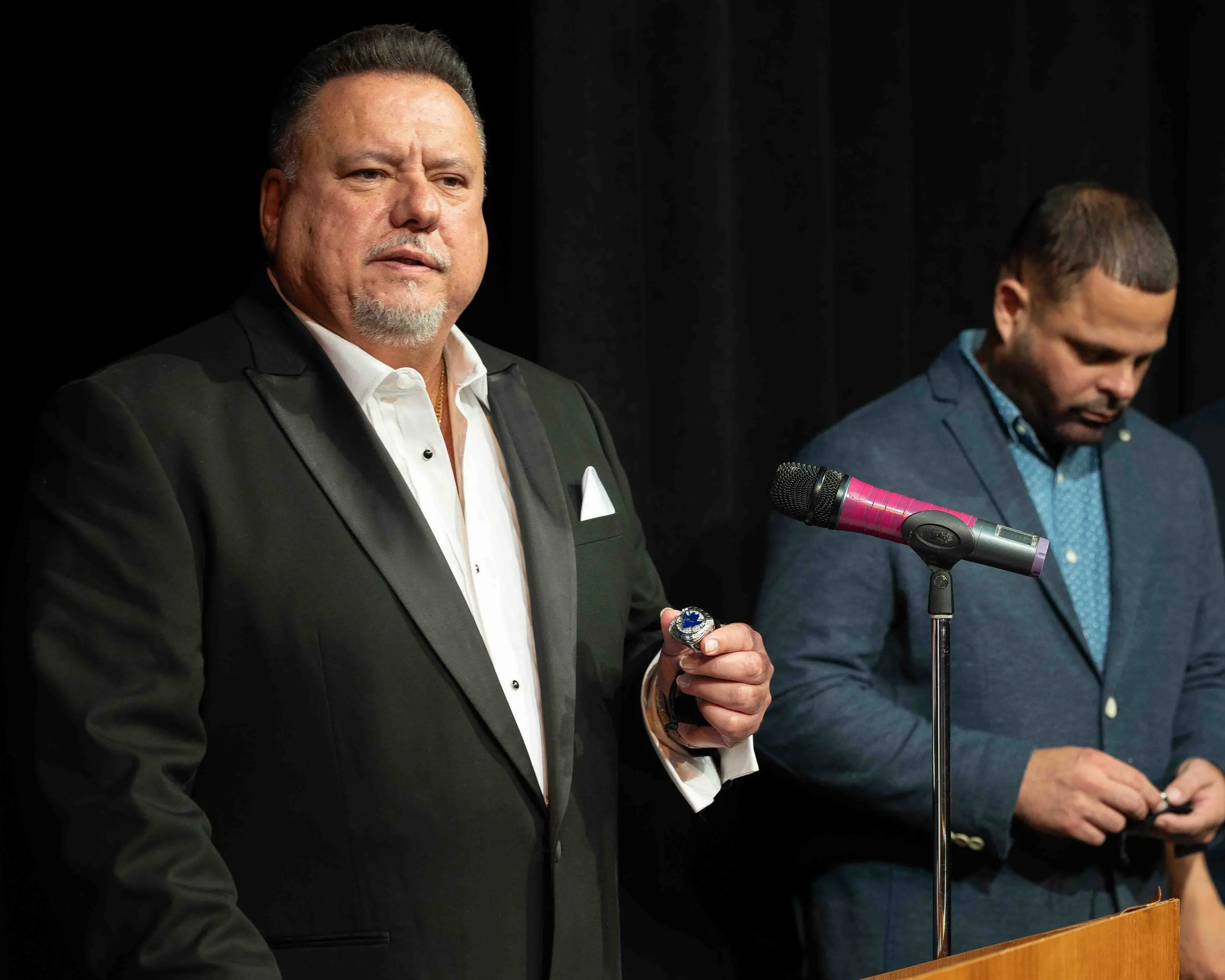 ABF's Art 'Pilin' Alvarez stands at a podium at an awards ceremony with basketball coach Macho Torres, in a blue blazer looking down at his phone behind him against a black background.