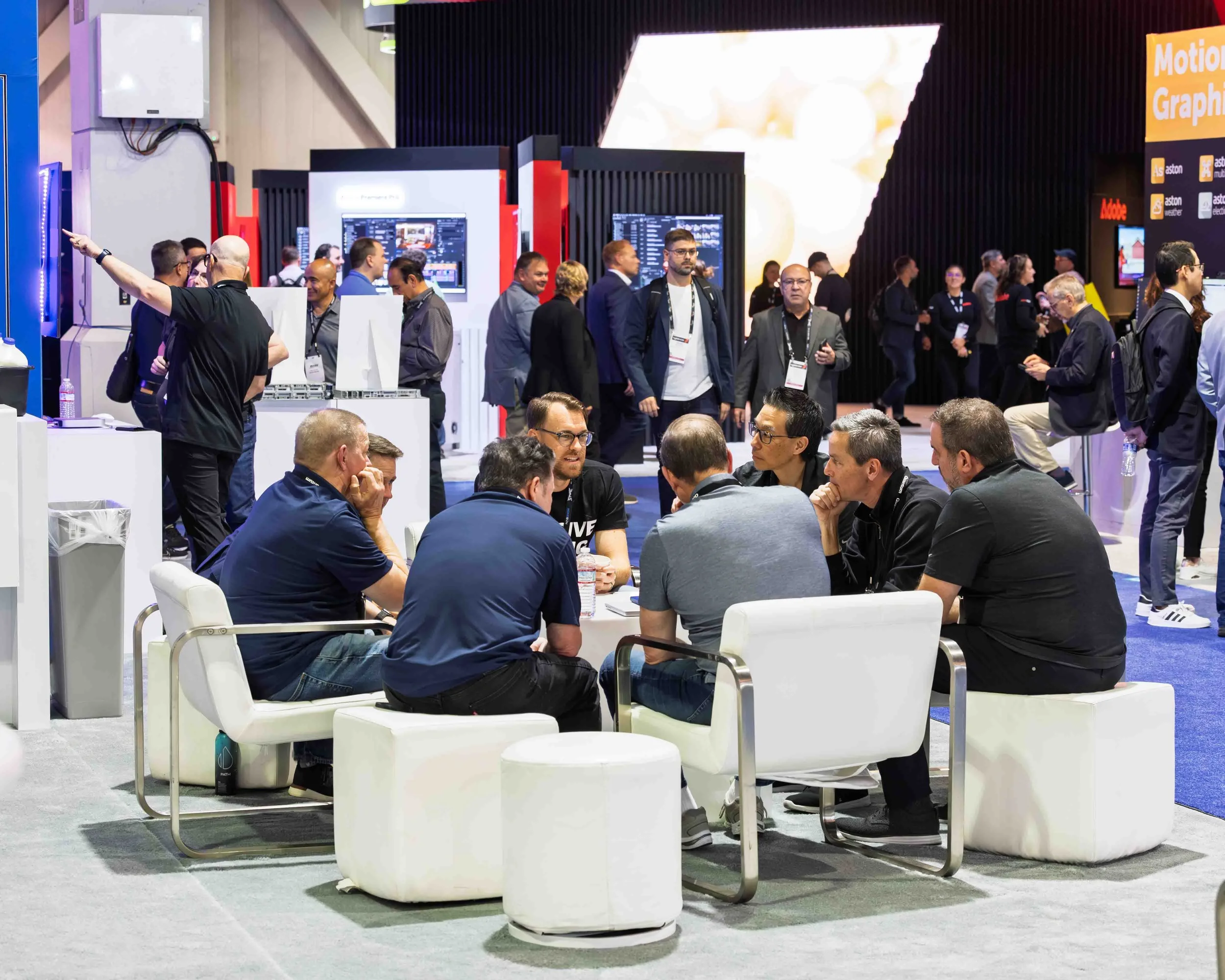 Group of people seated around a white table engaging in conversation at a corporate event or conference. Background shows various display booths and attendees walking around.