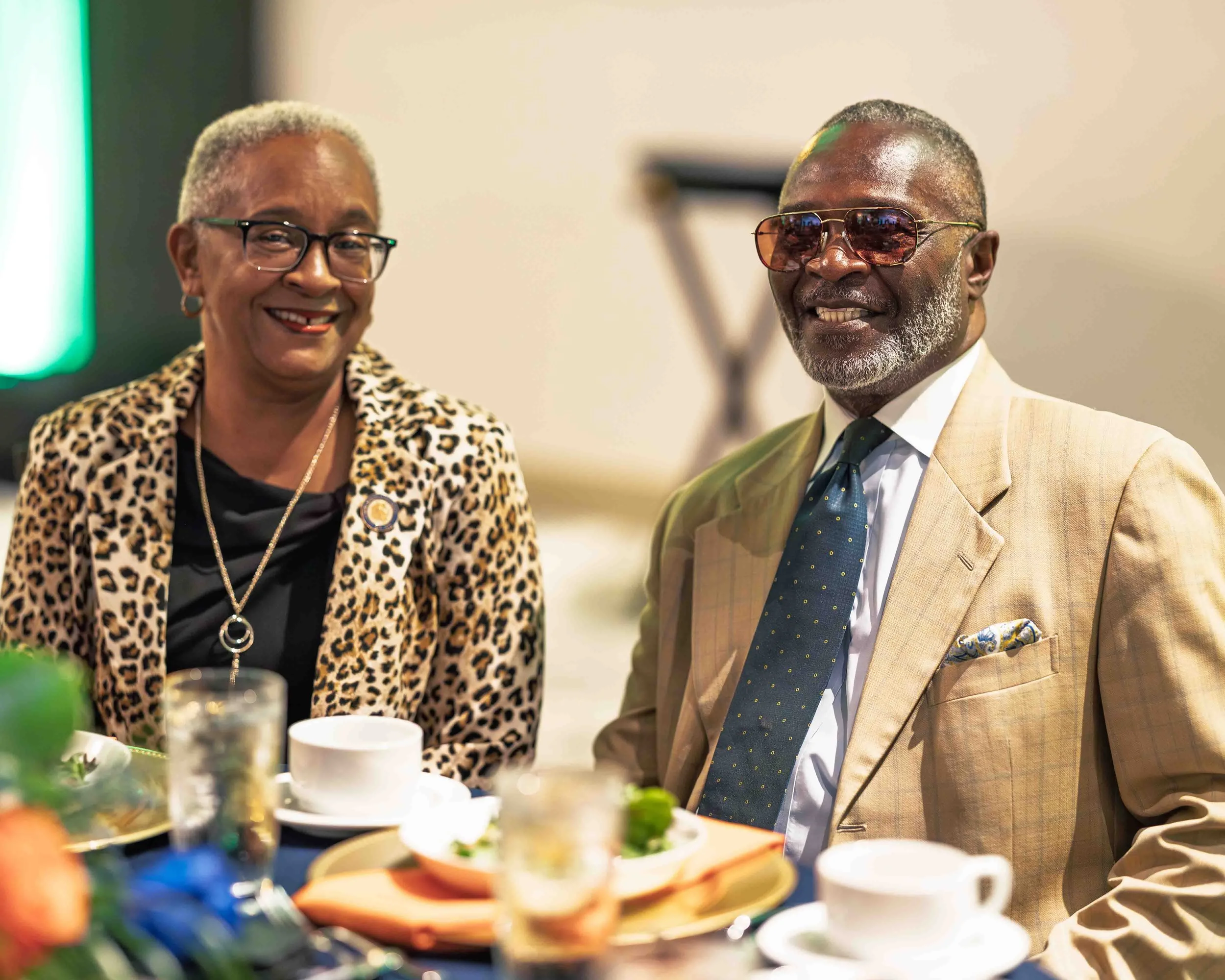 An older woman and man sitting at a dining table, smiling, in a well-lit indoor setting. The woman wears glasses, a leopard-print jacket, and a black top, with jewelry. The man wears glasses, a beige suit, white shirt, patterned tie, and pocket squar
