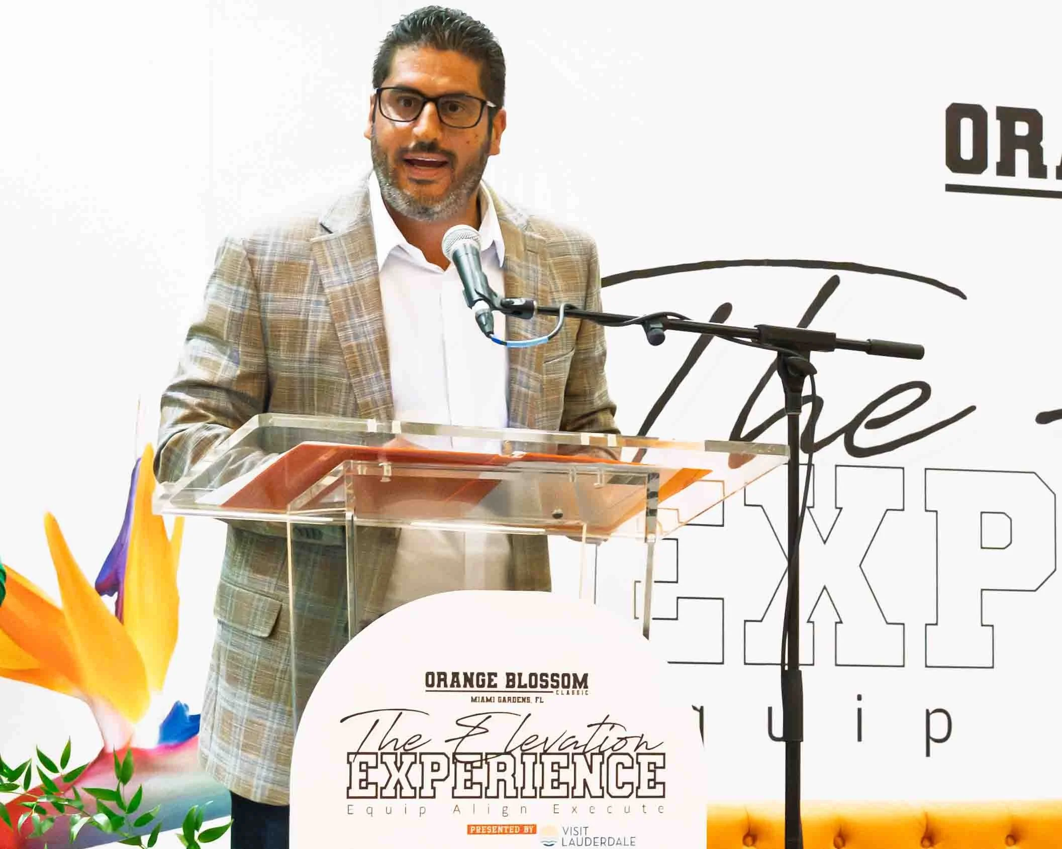 A man in a checkered blazer speaking at a podium with a microphone, with a colorful flower arrangement to his left. Background has text related to a corporate event called 'The Elevation Experience'.