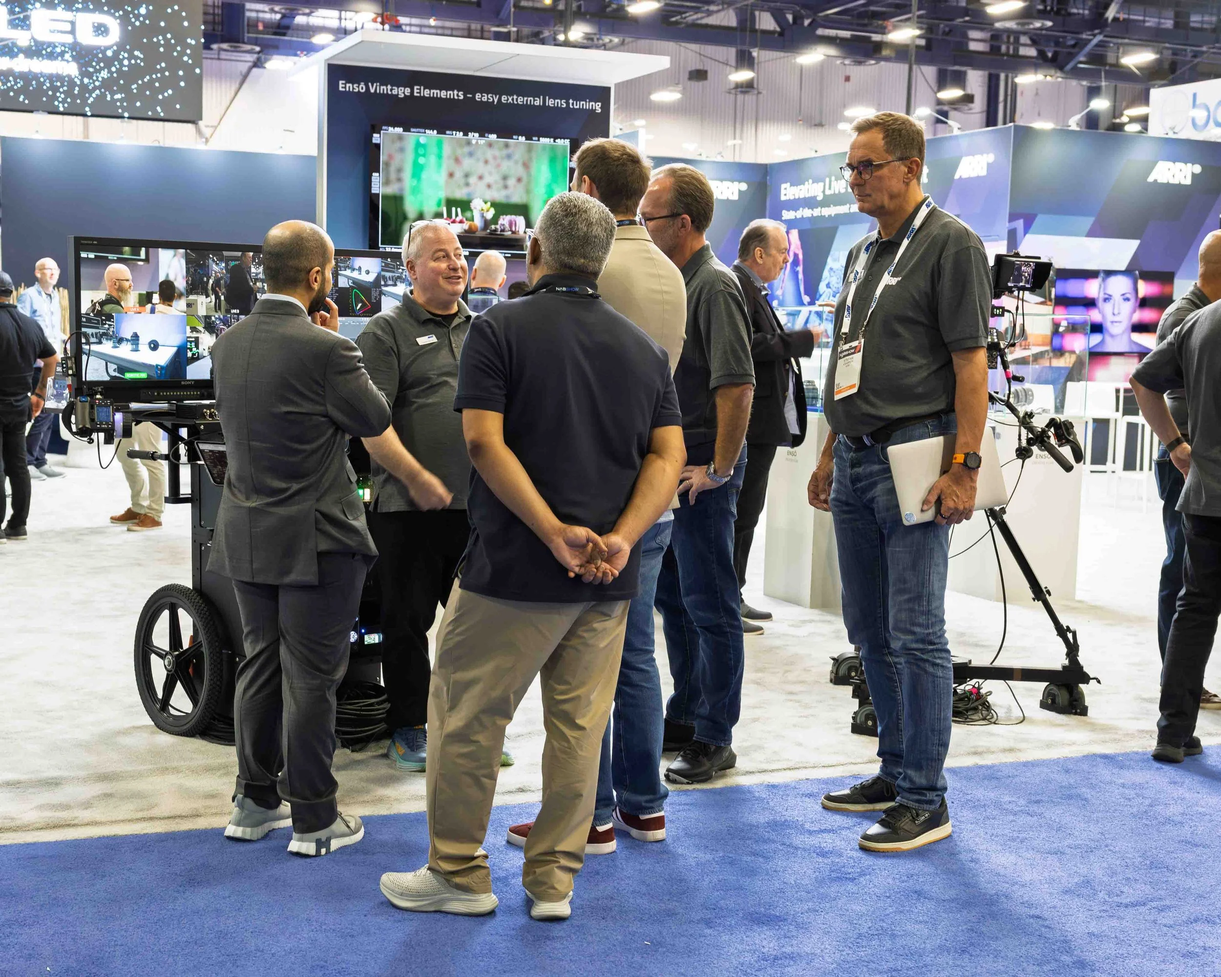 Group of people engaging in conversation at a technology trade show or conference with monitors and equipment displayed around them.