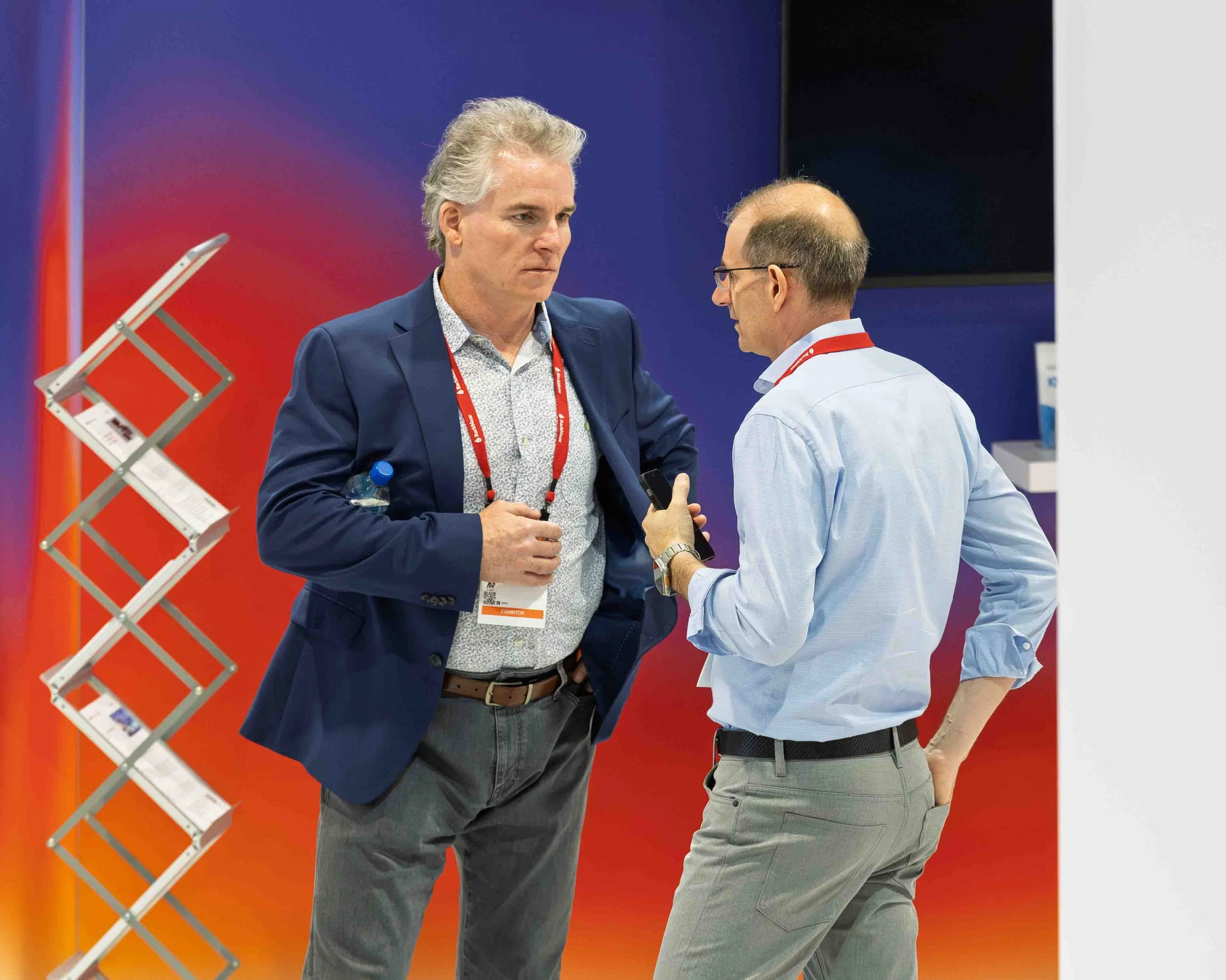 Two men engaged in conversation at a corporate event, standing near a display with a purple and orange background and a collapsible stand holding brochures.