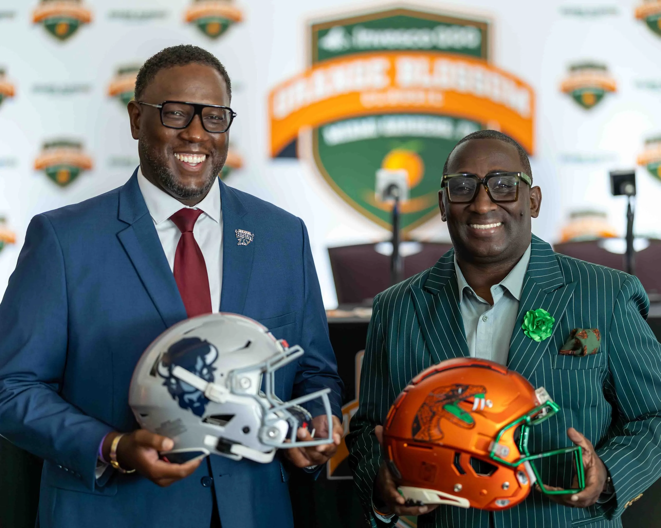 Howard University football coach Larry Scott and Florida A&M Football Coach James Colzie III in suits holding football helmets at a sports event, with a backdrop featuring a logo for the Detroit Lions at a press conference
