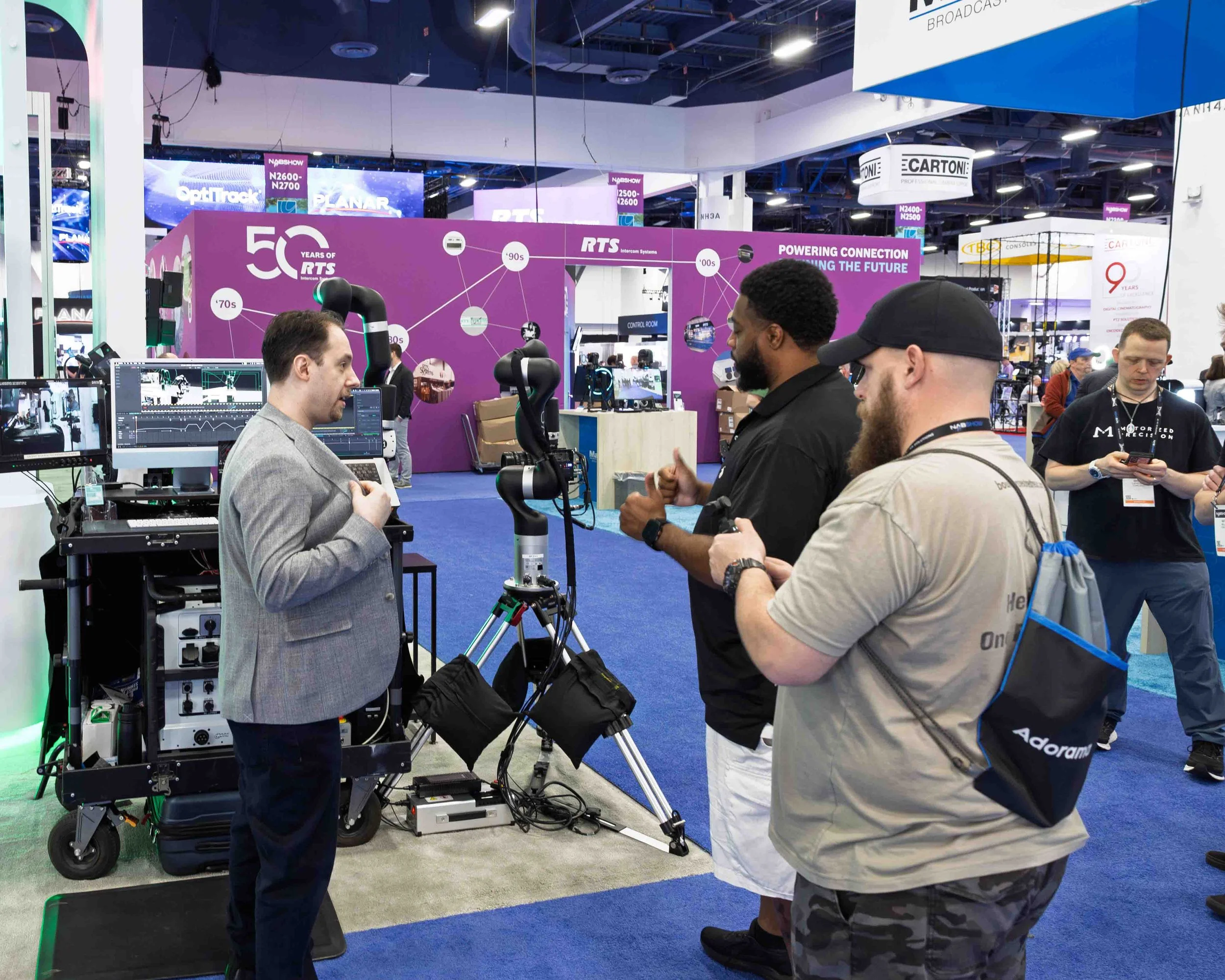 A presenter explaining technology to two attendees at a trade show booth, with computers, robotic arms, and screens displaying data around them.