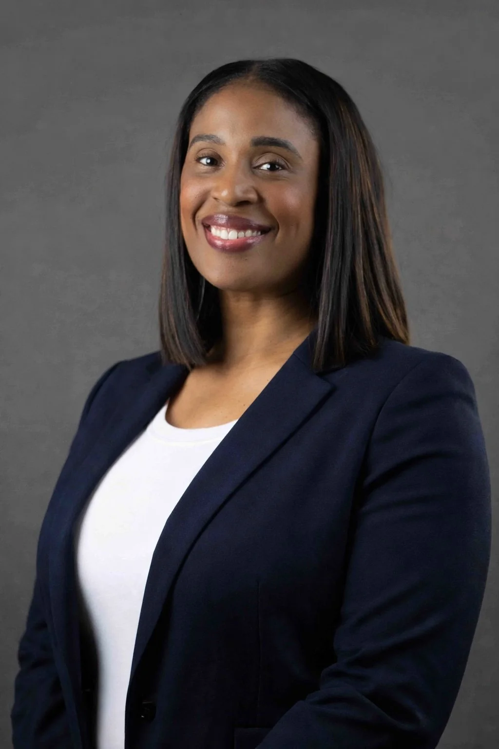 Headshot of a professional woman with shoulder-length dark hair smiling at the camera, wearing a navy blazer and white top against a gray background.