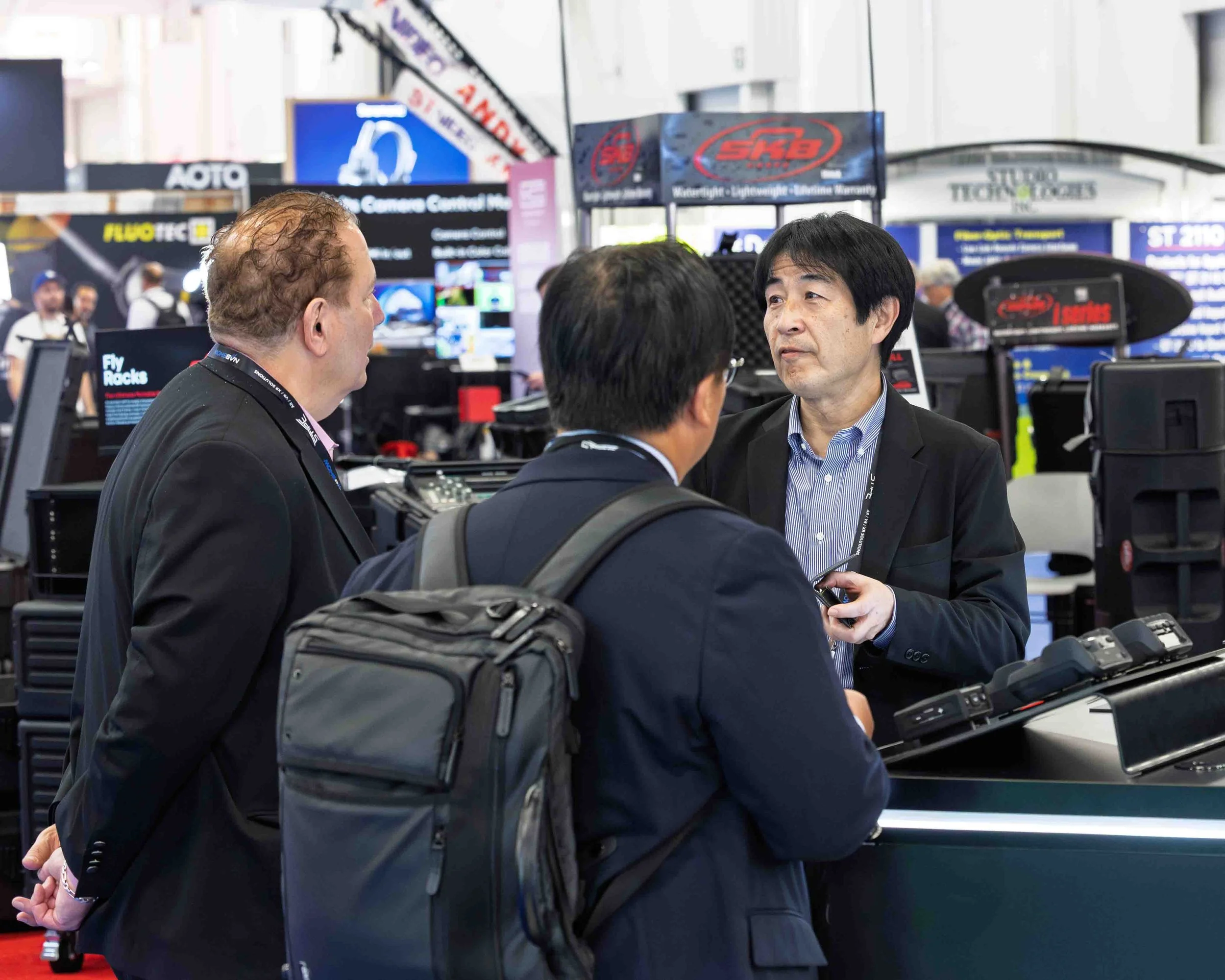 Three men in business attire engaged in conversation at a trade show or conference, with booths and electronic equipment in the background.