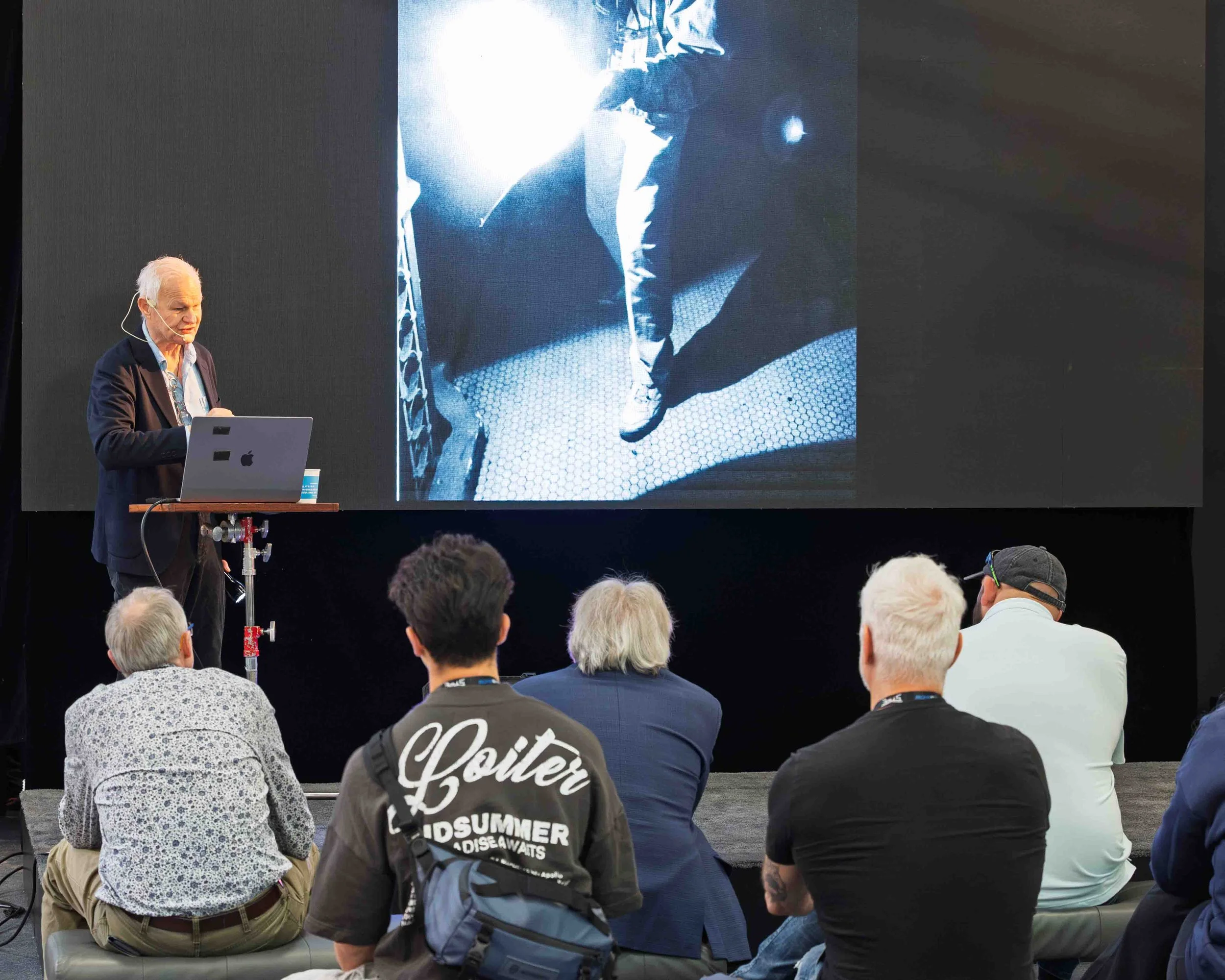 A man giving a presentation on an event stage with a large screen behind him showing an abstract black and white photo of a person walking. The audience is seated in front, attentively watching. The presenter is using a laptop.