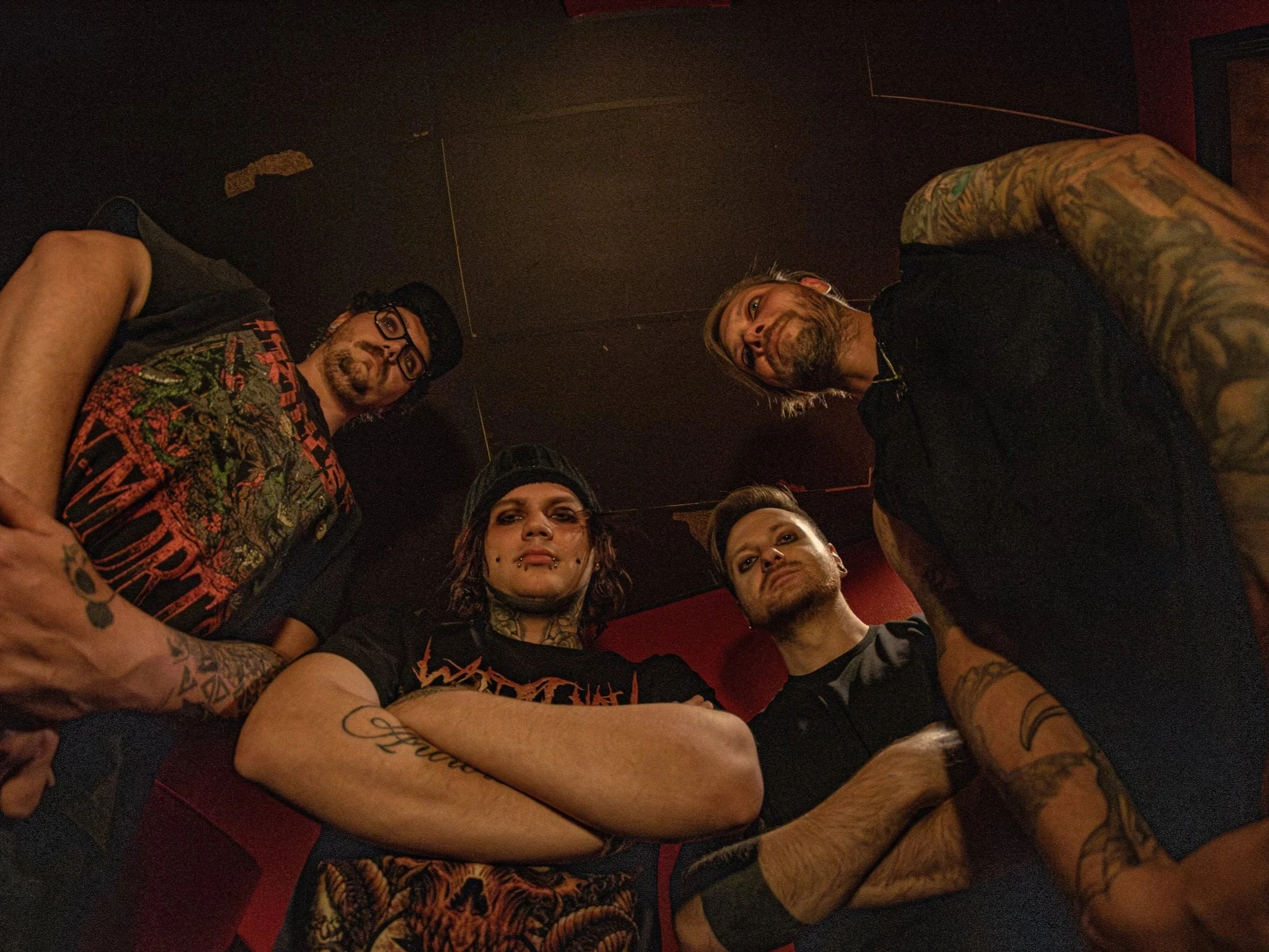 Five men with tattoos and piercings standing in a circle, looking down at the camera, in a dimly lit room.