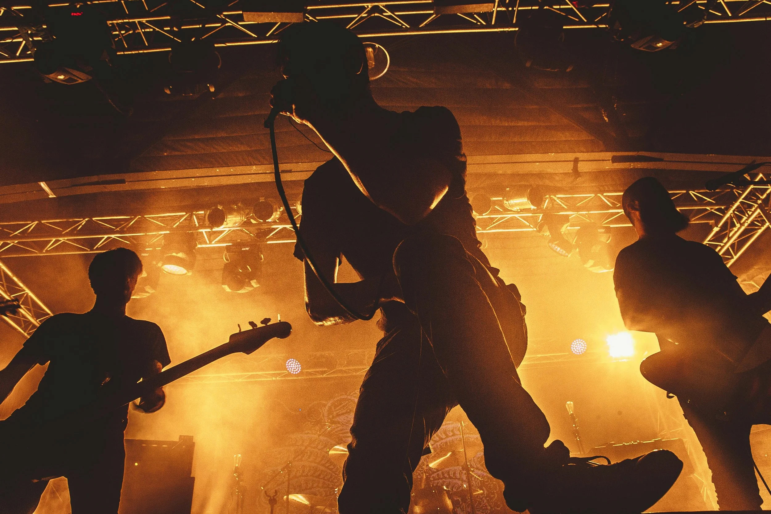 A musician playing an electric guitar on stage, with drums and amplifiers in the background, under stage lighting.