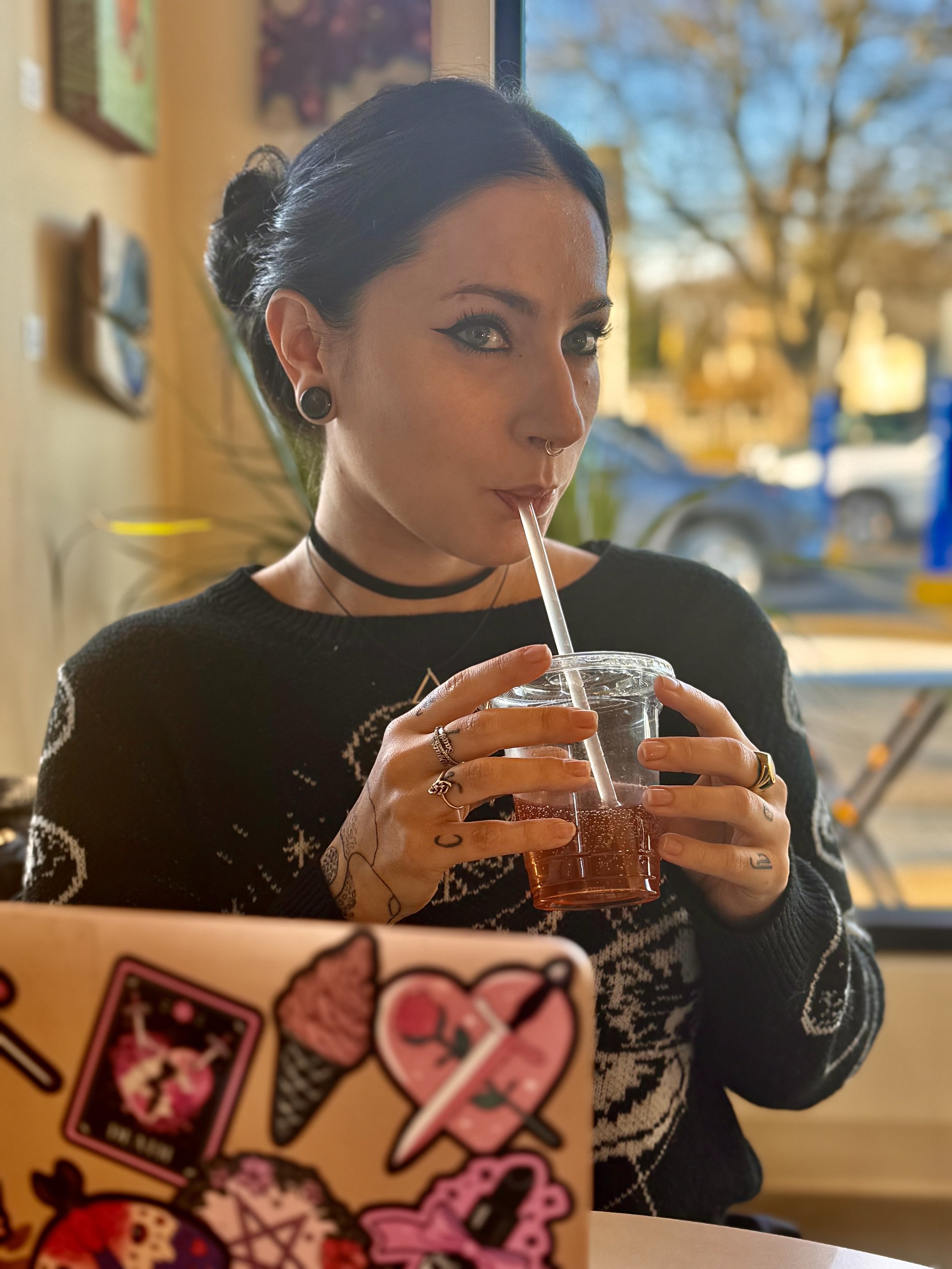 A woman with black hair styled in a bun, wearing a black sweater with white patterns, and a black choker, drinks a pink beverage with a straw in a cafe or restaurant during daylight. She has piercings and tattoos visible on her hands.