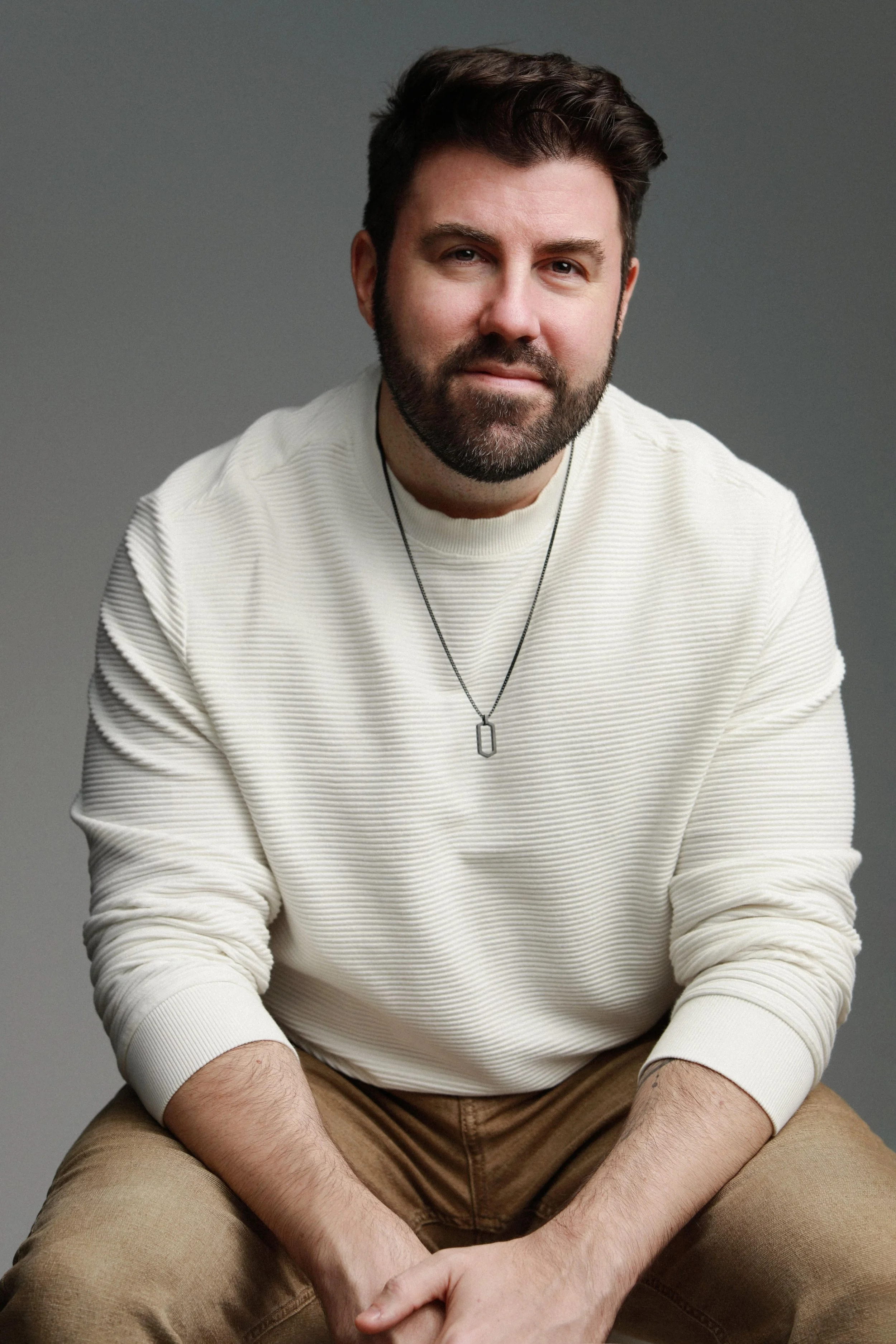 A man with dark hair and a beard wearing a cream-colored striped long sleeve shirt, a necklace with a rectangular pendant, and khaki pants, sitting against a gray background.