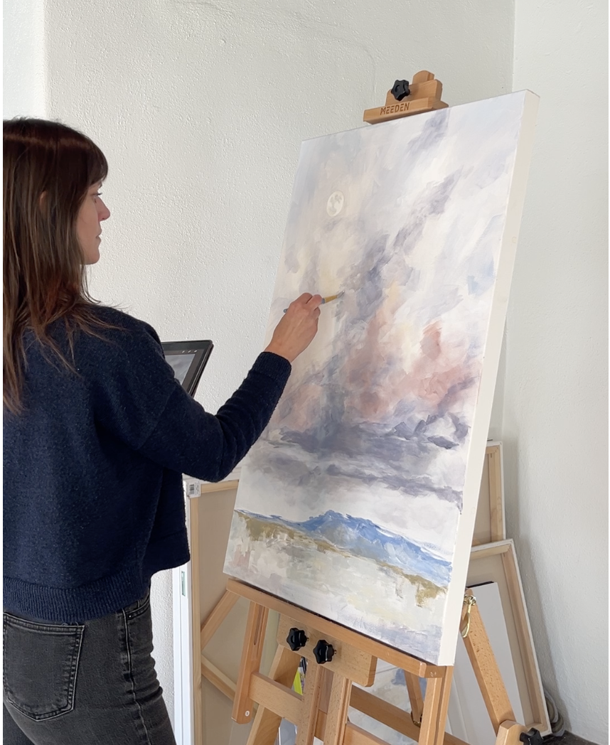 A woman painting on a large canvas set up on an easel, using a brush on a partly cloudy sky with mountains and a body of water below.