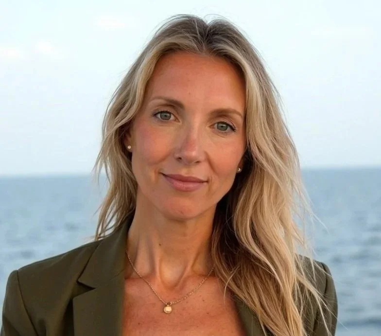 A woman with long blonde hair, wearing a green blazer and a gold necklace, standing outdoors near the ocean with a cloudy sky in the background. Bethesda psychiatrist.