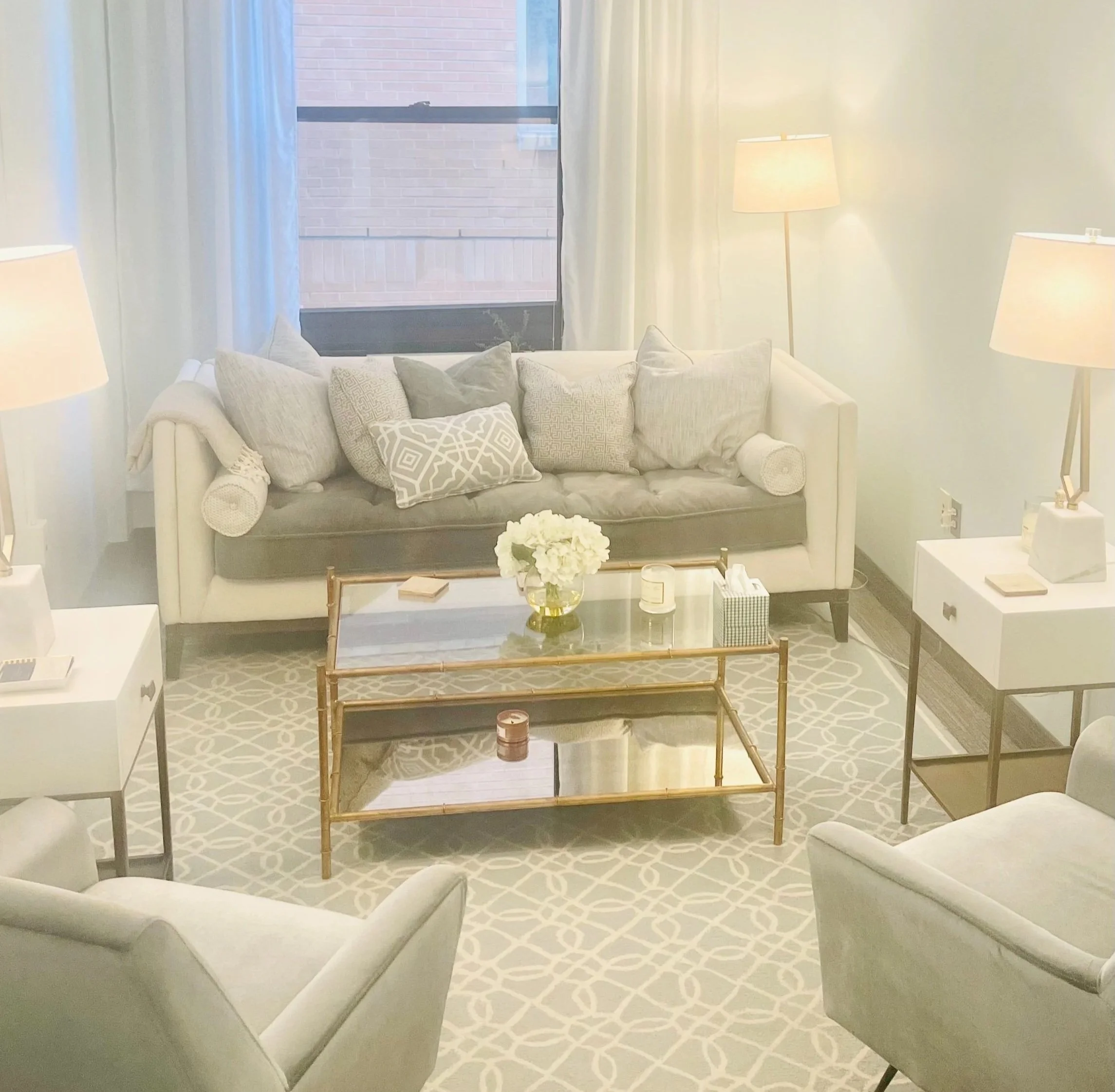 A cozy living room with a white sofa, multiple pillows, and two side tables with lamps, a coffee table with a flower vase, candles, and decorative items, and an armchair, all on a patterned rug near a window with curtains. chevy chase psychiatry