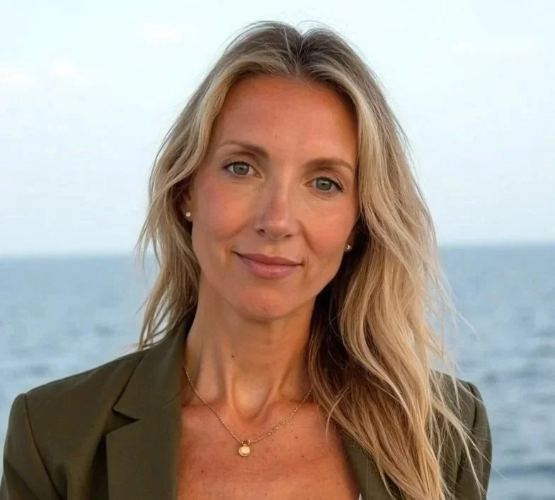 A blonde woman with wavy hair smiling at the camera outdoors by the ocean, wearing a green blazer and a gold necklace. NOVA psychiatrist