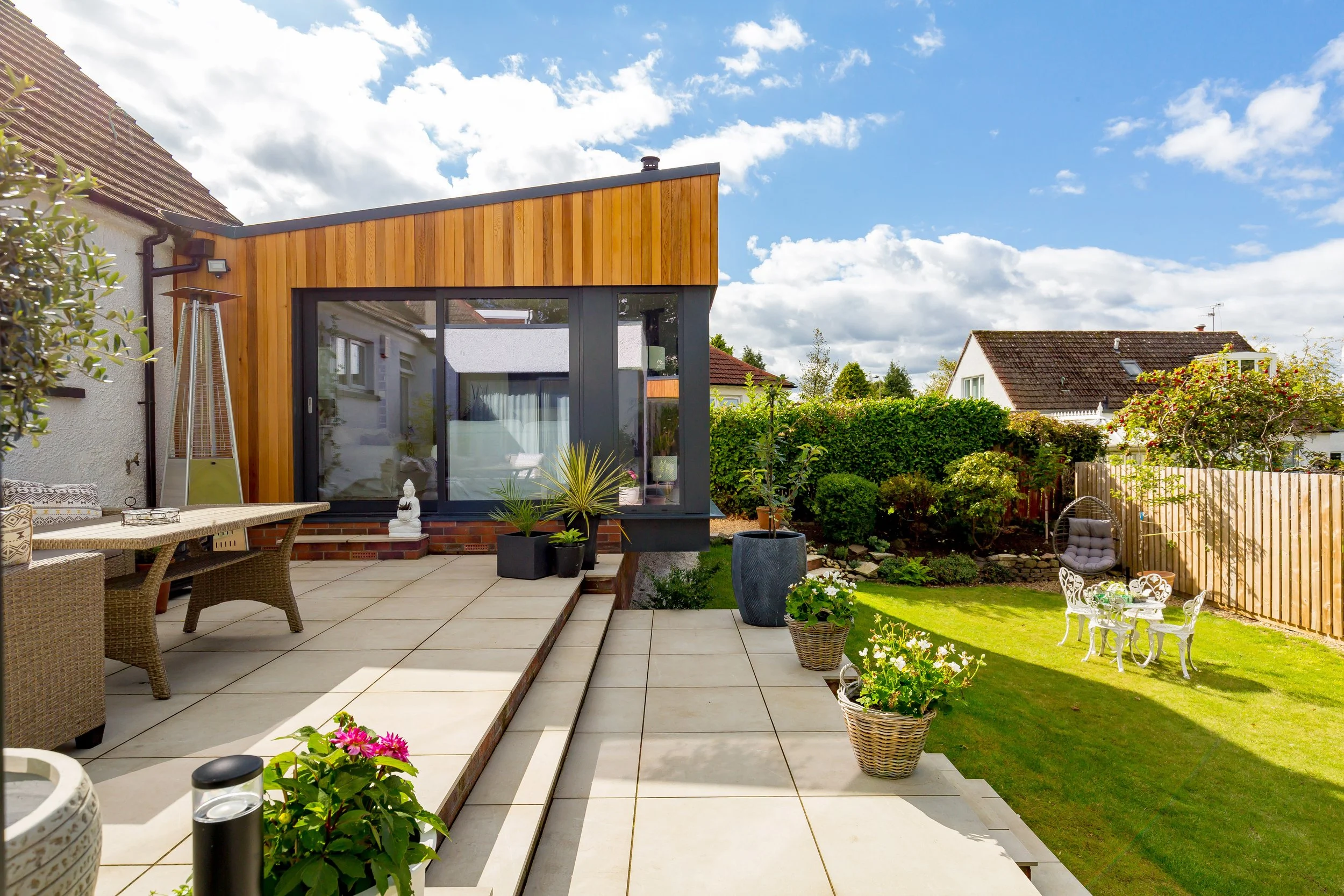 Backyard patio with outdoor furniture, potted plants, and a modern extension of a house with wood and glass surfaces under a partly cloudy sky.