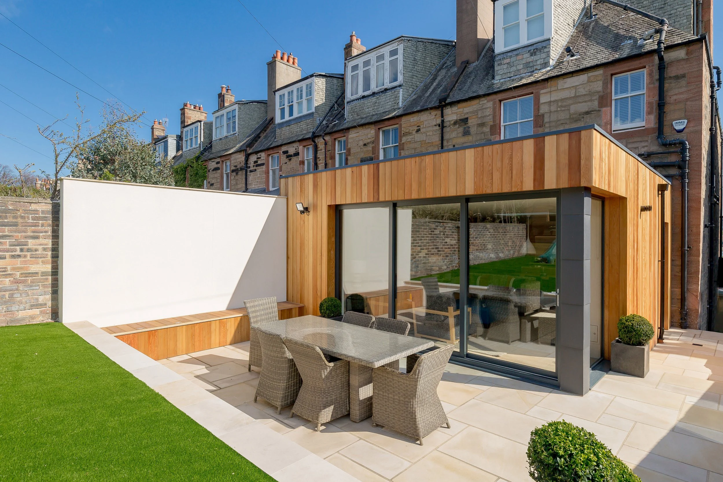 Modern backyard patio with outdoor dining area, wicker chairs, glass sliding doors, wooden exterior wall, small green plants, trimmed bushes, stone patio, and a white privacy wall, with old brick townhouses in the background under a clear blue sky.