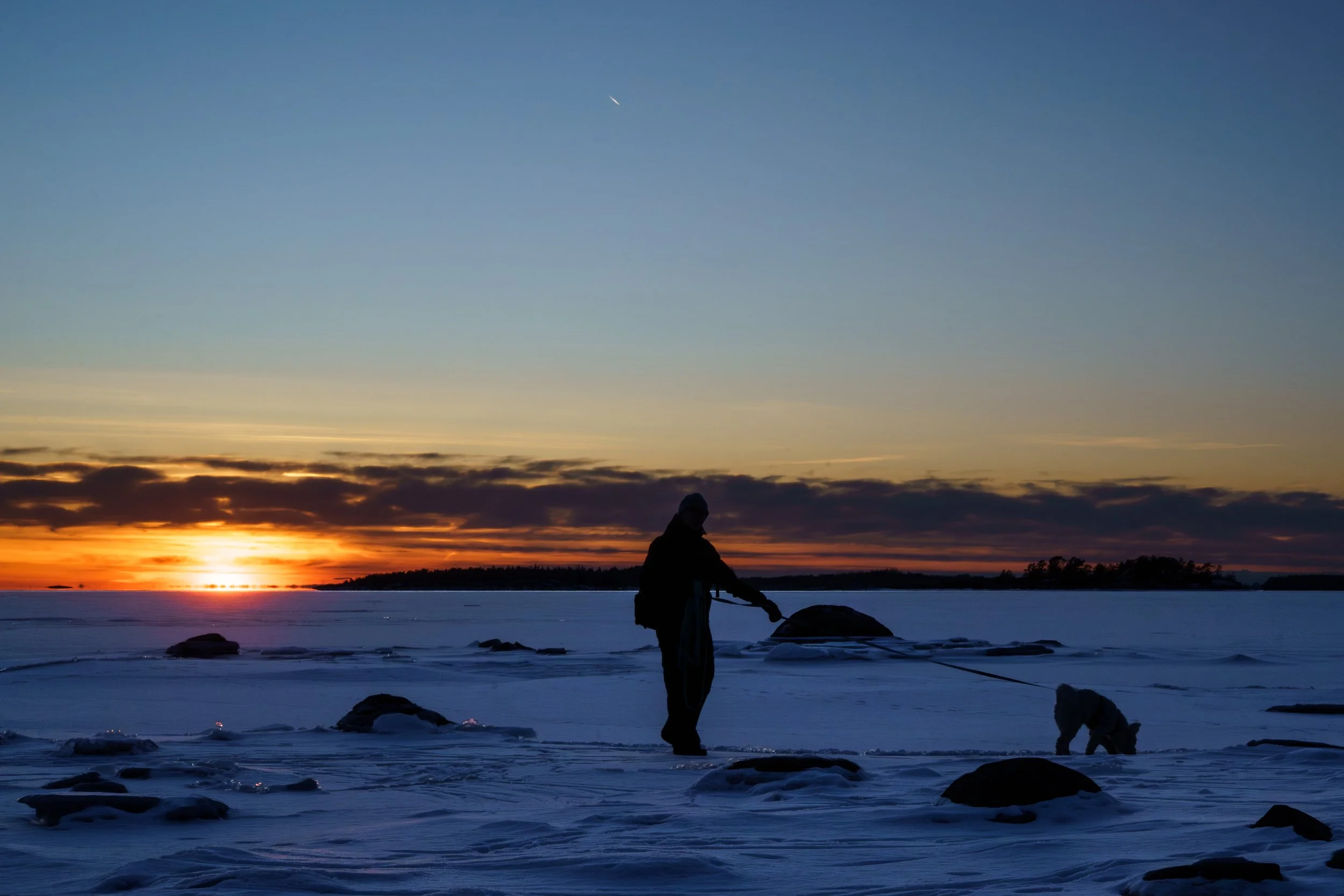 Person walking a dog across a snowy landscape at sunset.