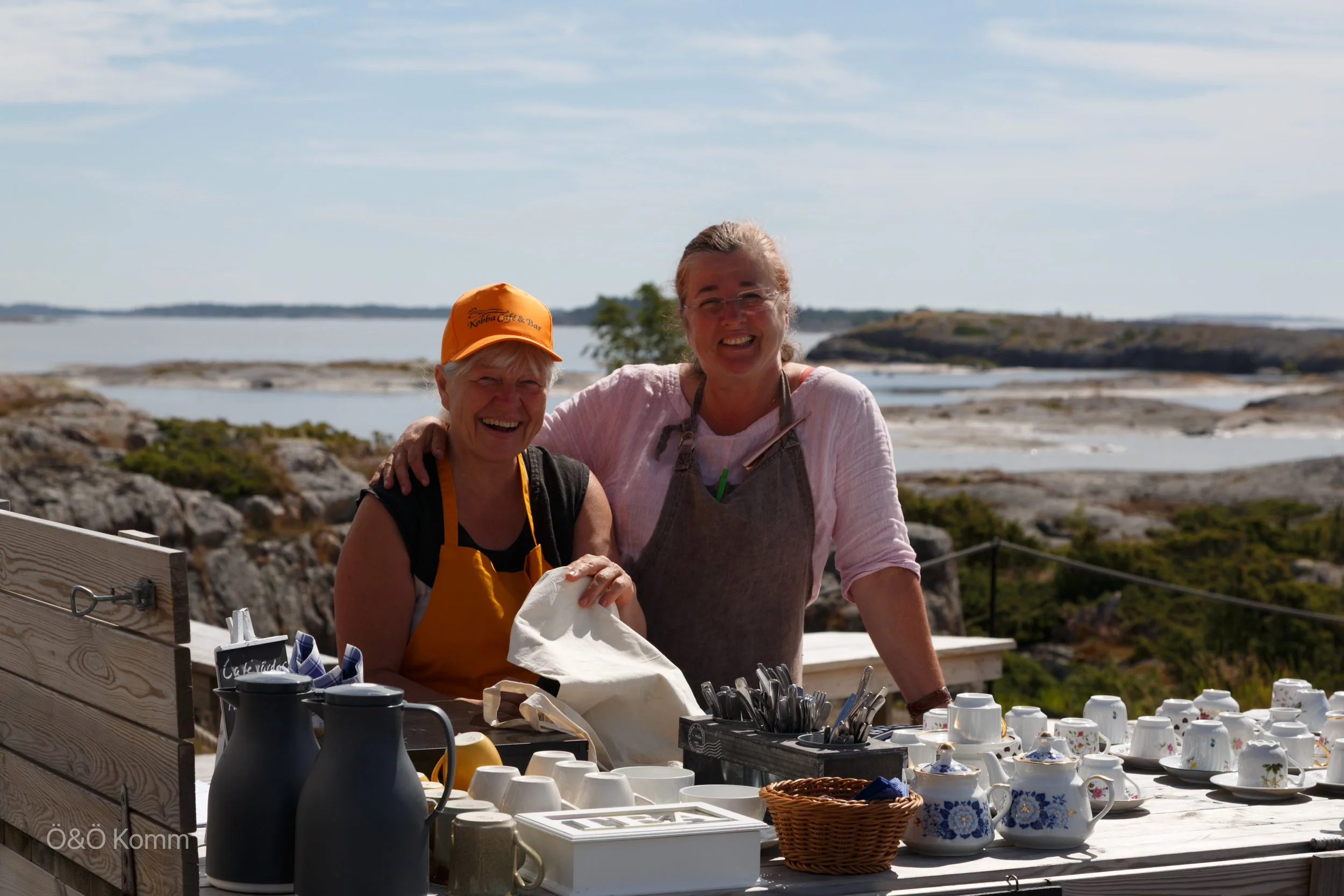 Two women smiling at a table with various dishes and cups outdoors near a body of water.
