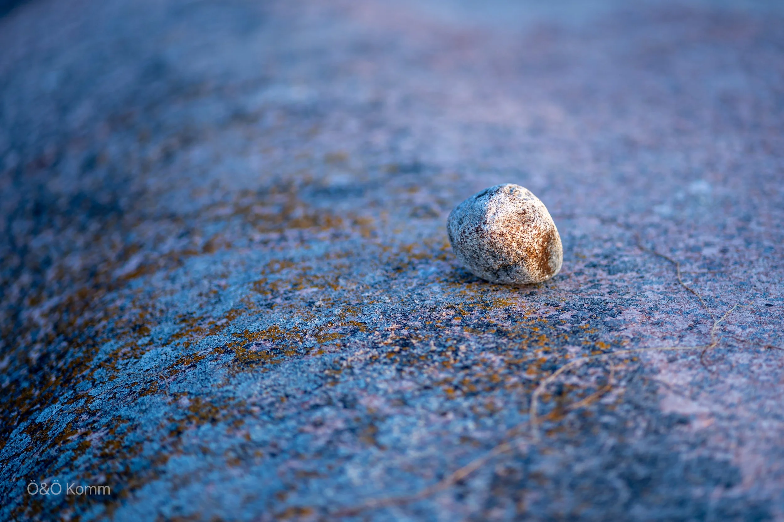 Close-up of a small, rounded stone resting on a sandy surface with a mix of blue, brown, and pink hues.