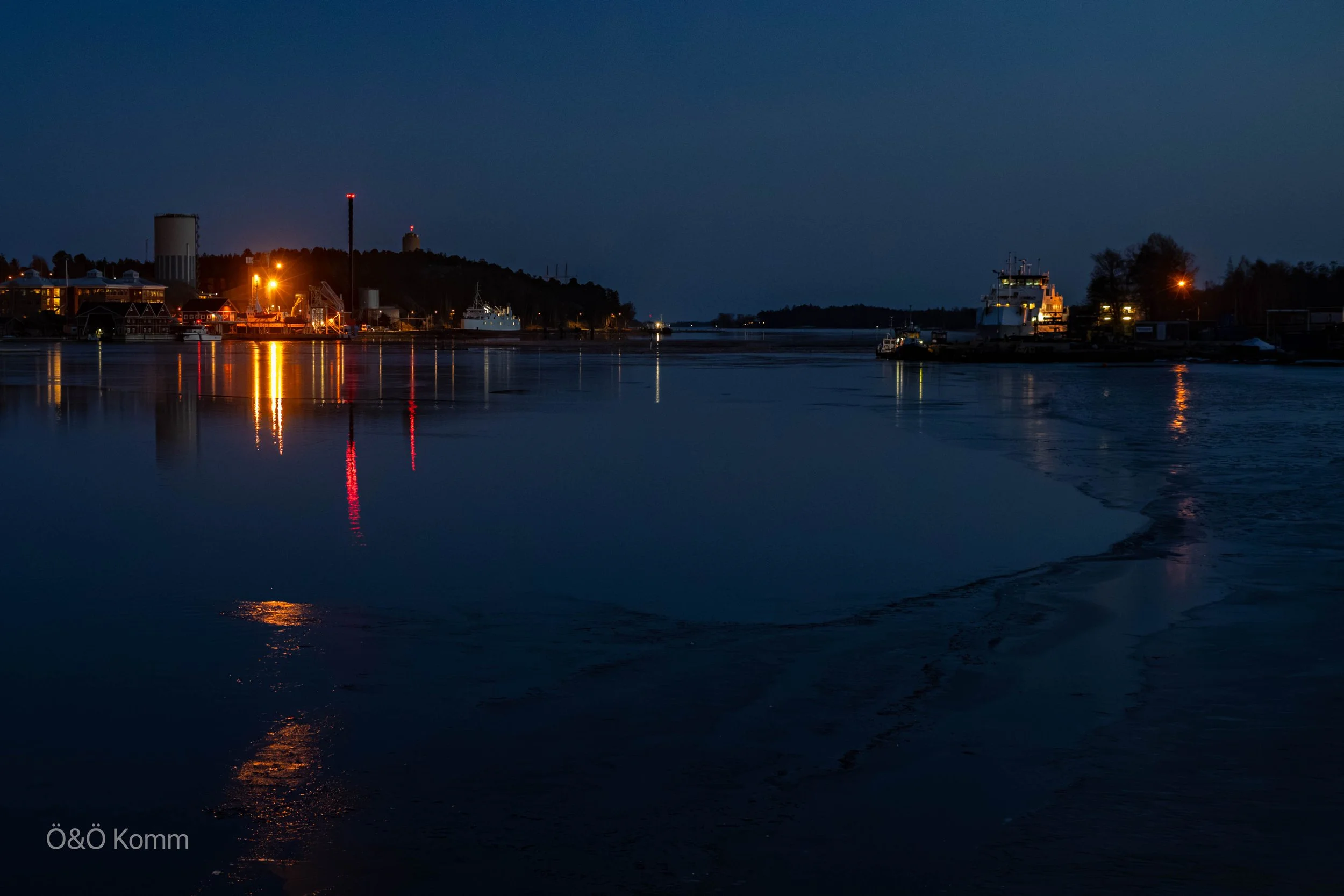 Nighttime photo of a harbor with boats, buildings, and distant hills, illuminated by streetlights and reflecting on calm water.