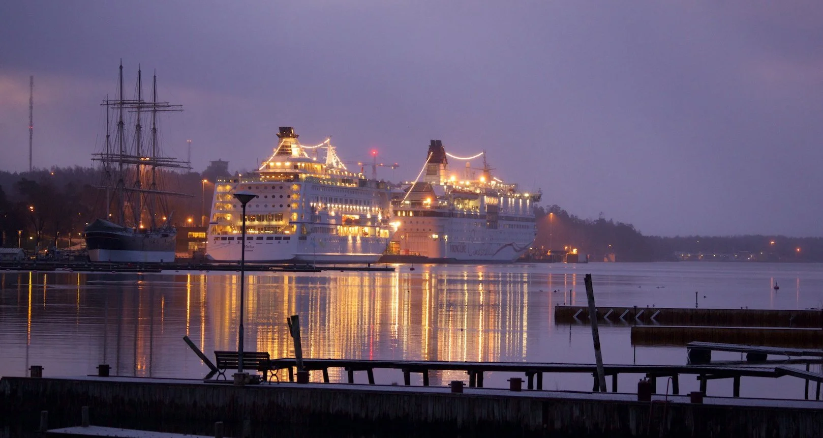 A large cruise ship is docked at a marina during twilight, with reflections on the calm water and a distant shoreline in the background.