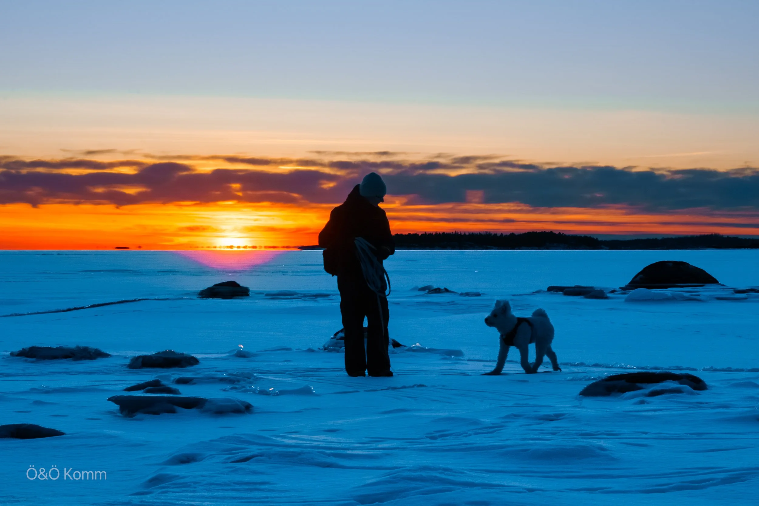 A person and a dog standing on a snowy landscape during sunset with colorful sky and clouds.