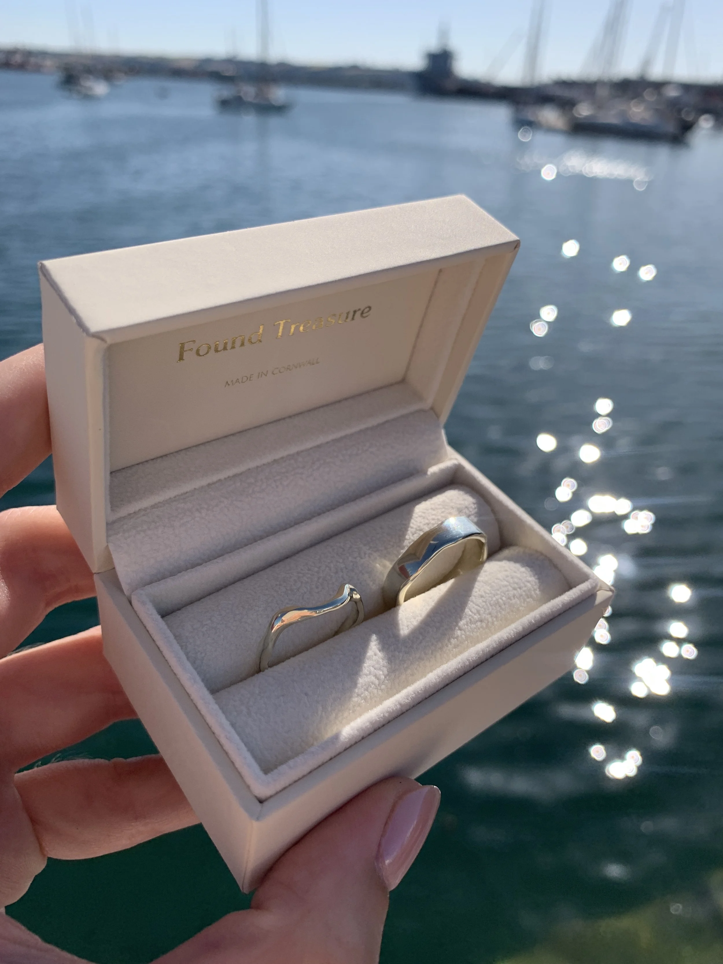 A pair of silver wedding rings in a white jewelry box with gold lettering, held outdoors near a harbor with boats and water in the background.