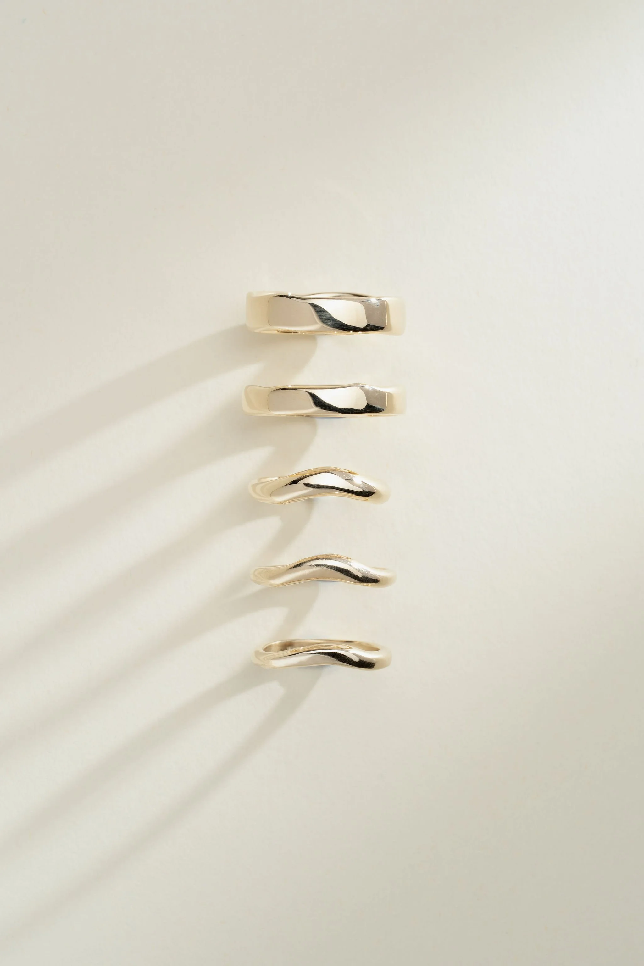Five gold rings with wavy and smooth designs, arranged vertically against a white background, casting shadows.