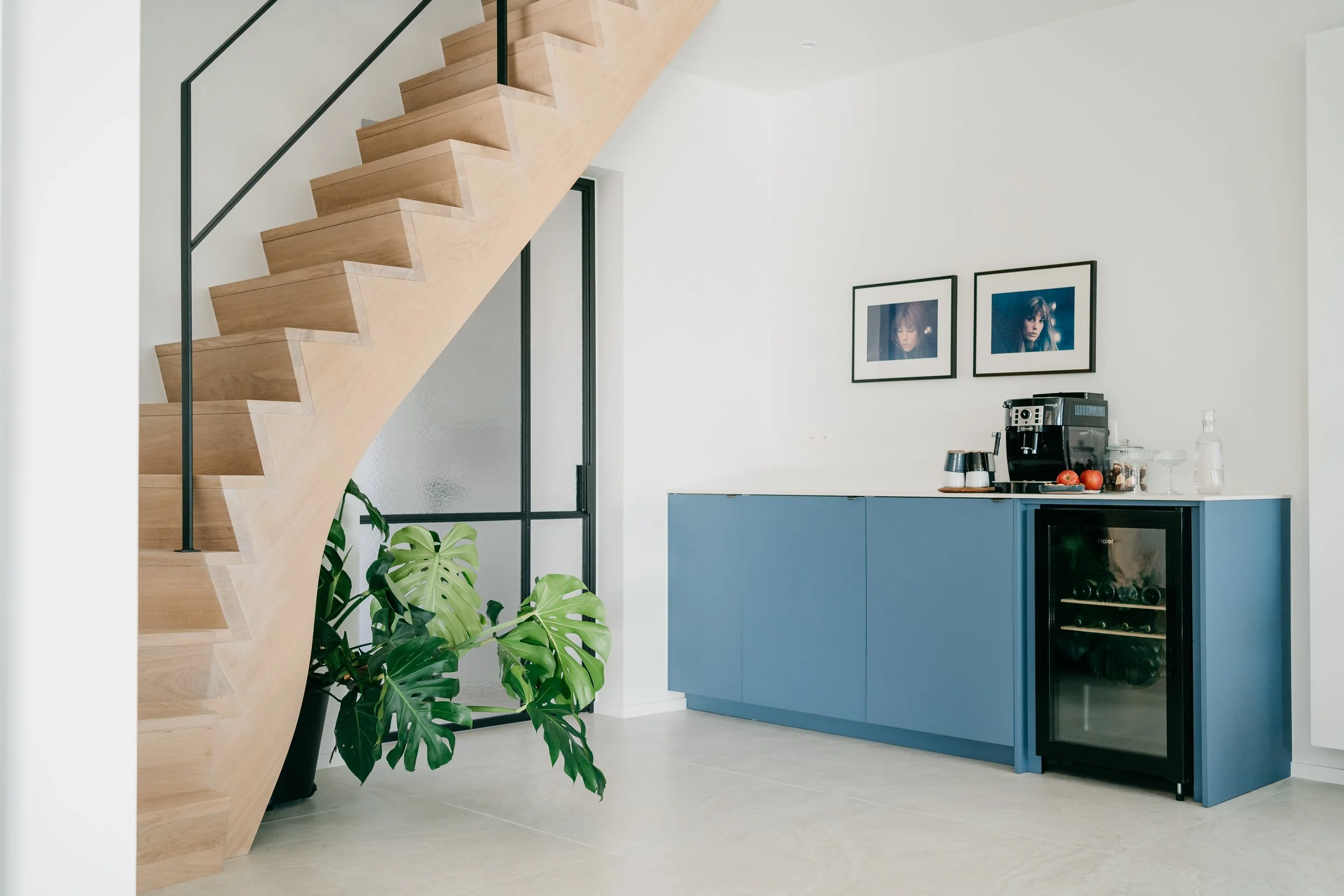 Interieur van een moderne keuken met een houten open trap, blauwe kasten, een koffiezetapparaat, drie appels, glazen en een wijnkoelkast.