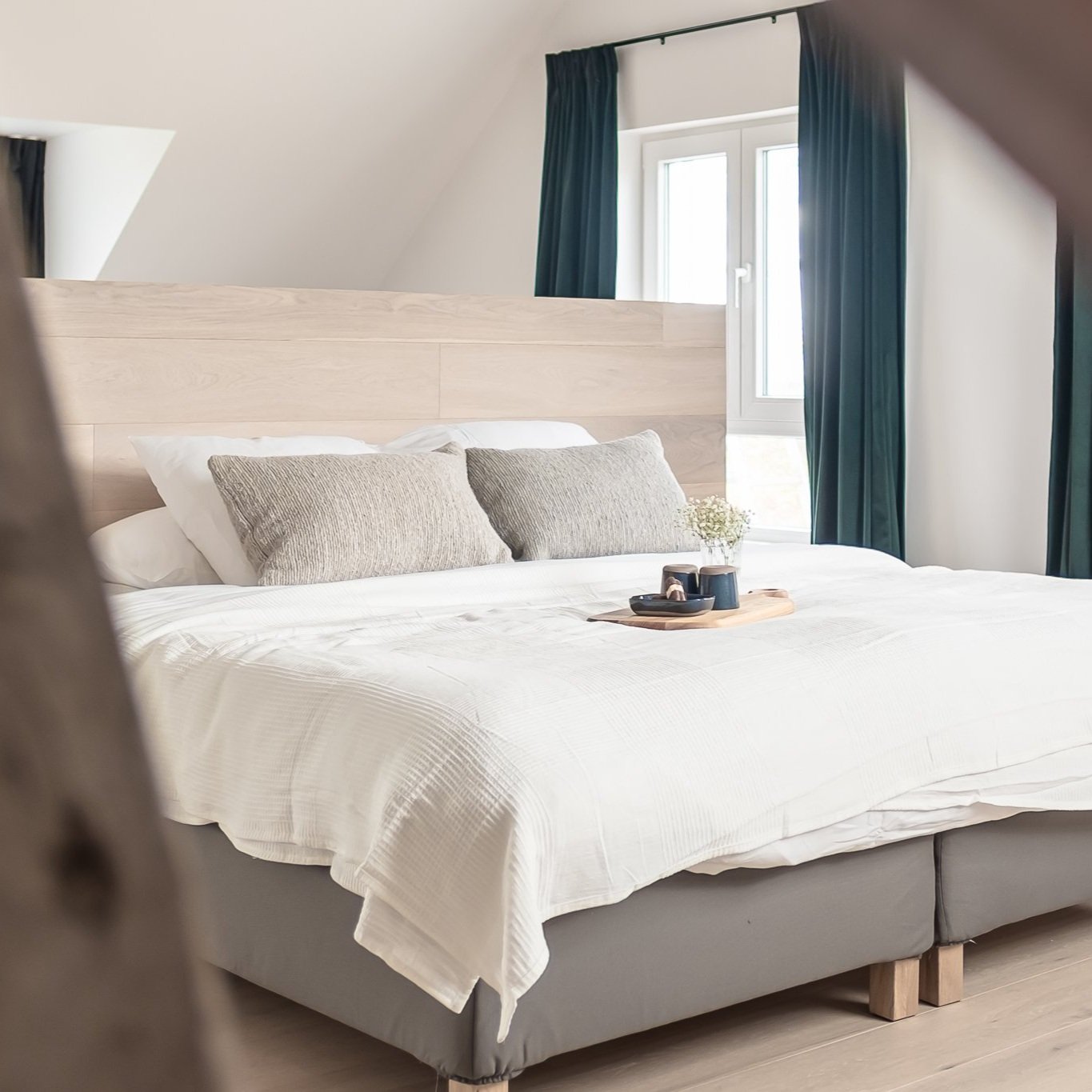 Een nette slaapkamer met een groot bed bedekt met een wit dekbed en twee grijze kussens. Op het bed ligt een houten dienblad met een vaas bloemen en twee bekers. Achter het bed bevinden zich grote ramen met blauwe gordijnen. Het plafond is schuinaardig, en de kamer heeft een rustige, lichte uitstraling.