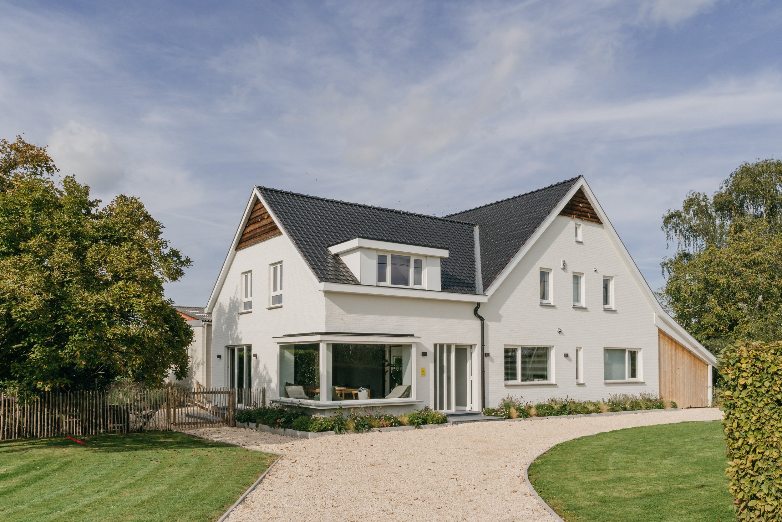 Moderne witte woning met een groene tuin en een grindpad voor de ingang, omringd door bomen en een hek onder een bewolkte blauwe hemel.