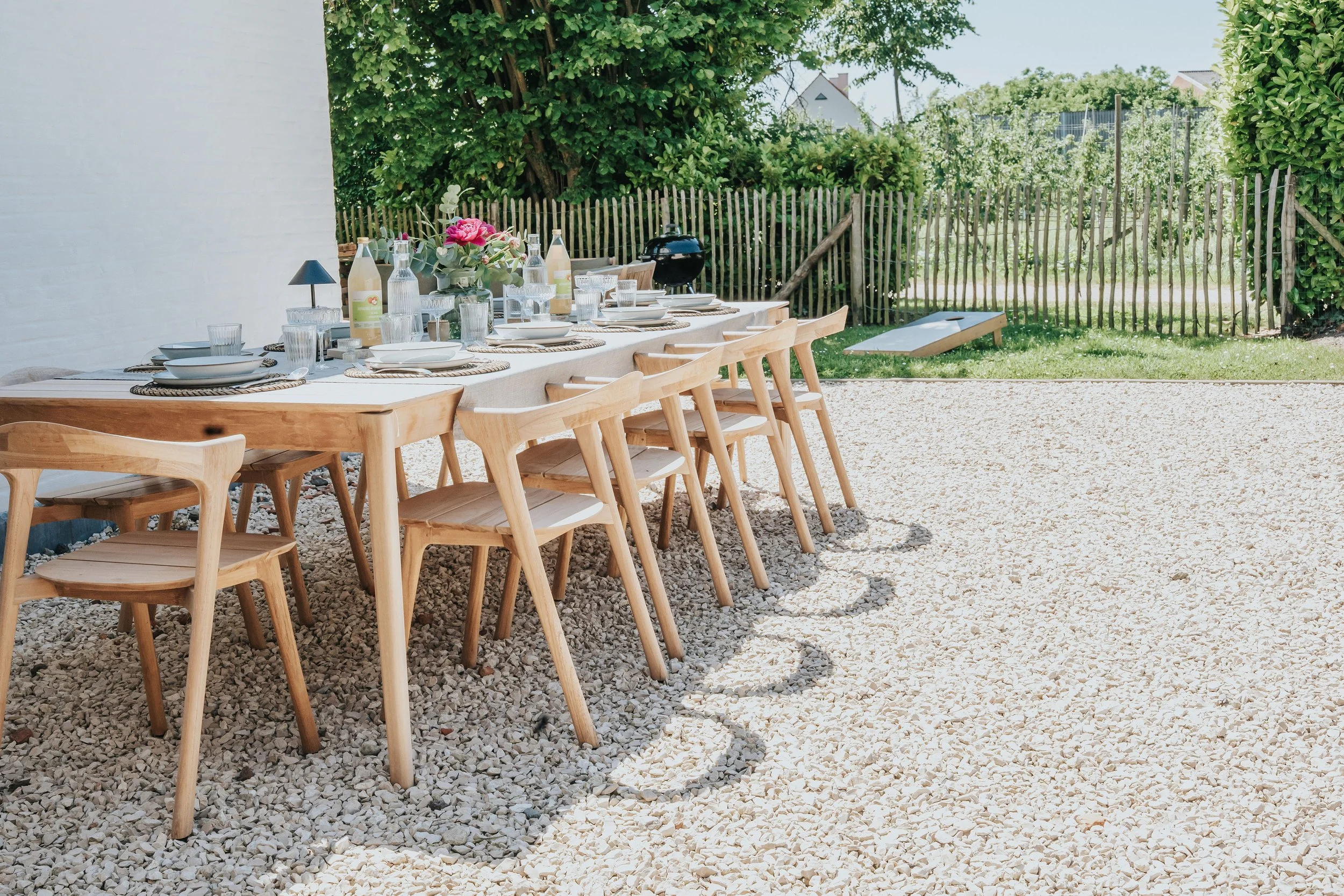 Eettafel met wit tafelkleed, servies, glazen, flesjes, bloemen, en een barbecue in een tuin met groene heggen en bomen.