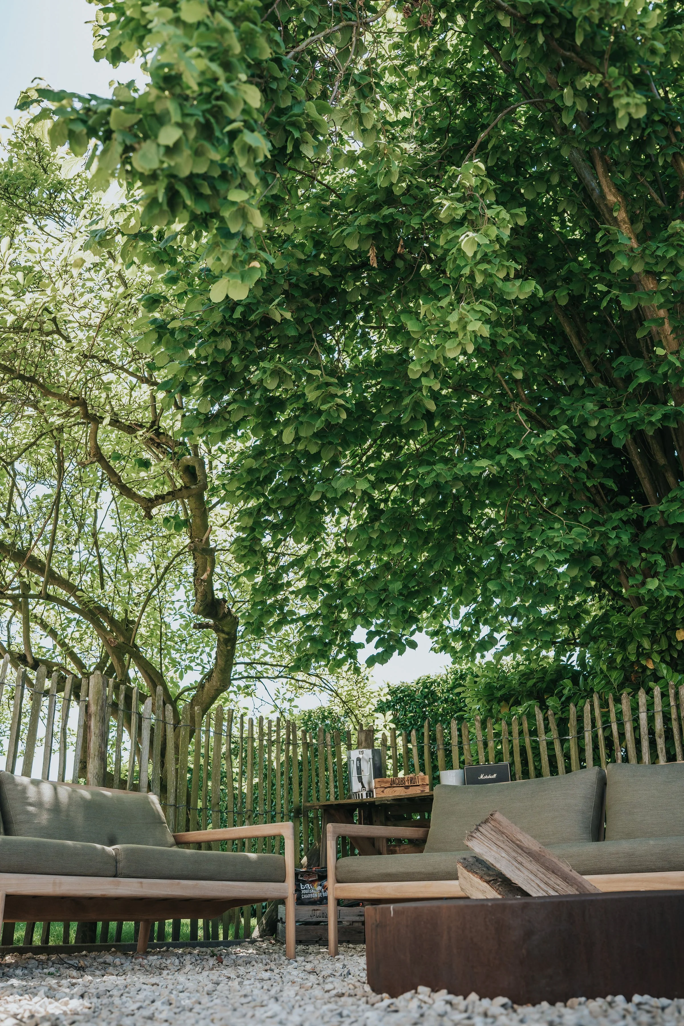 Buiten zitje met grijze bank, houten fauteuil en vuurkorf op grind onder een grote boom met groene bladeren, omgeven door een houten hek, op een zonnige dag.