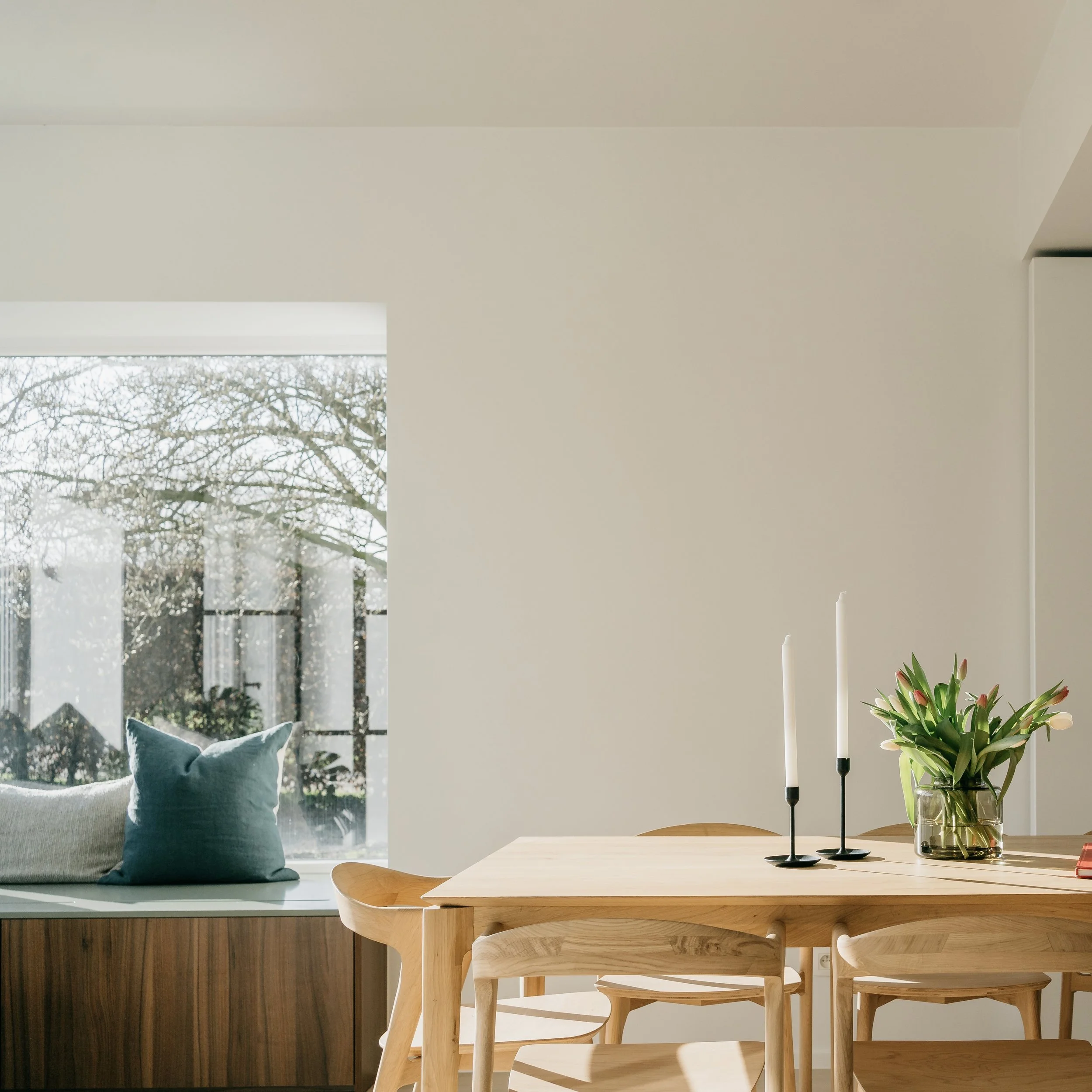 Image van een interieur met een eettafel en houten stoelen, een plant in een vaas, twee kandelaren met kaarsen, en een grote raamtoonbank met een stoel en een kussen bij een raam met uitzicht op bomen.