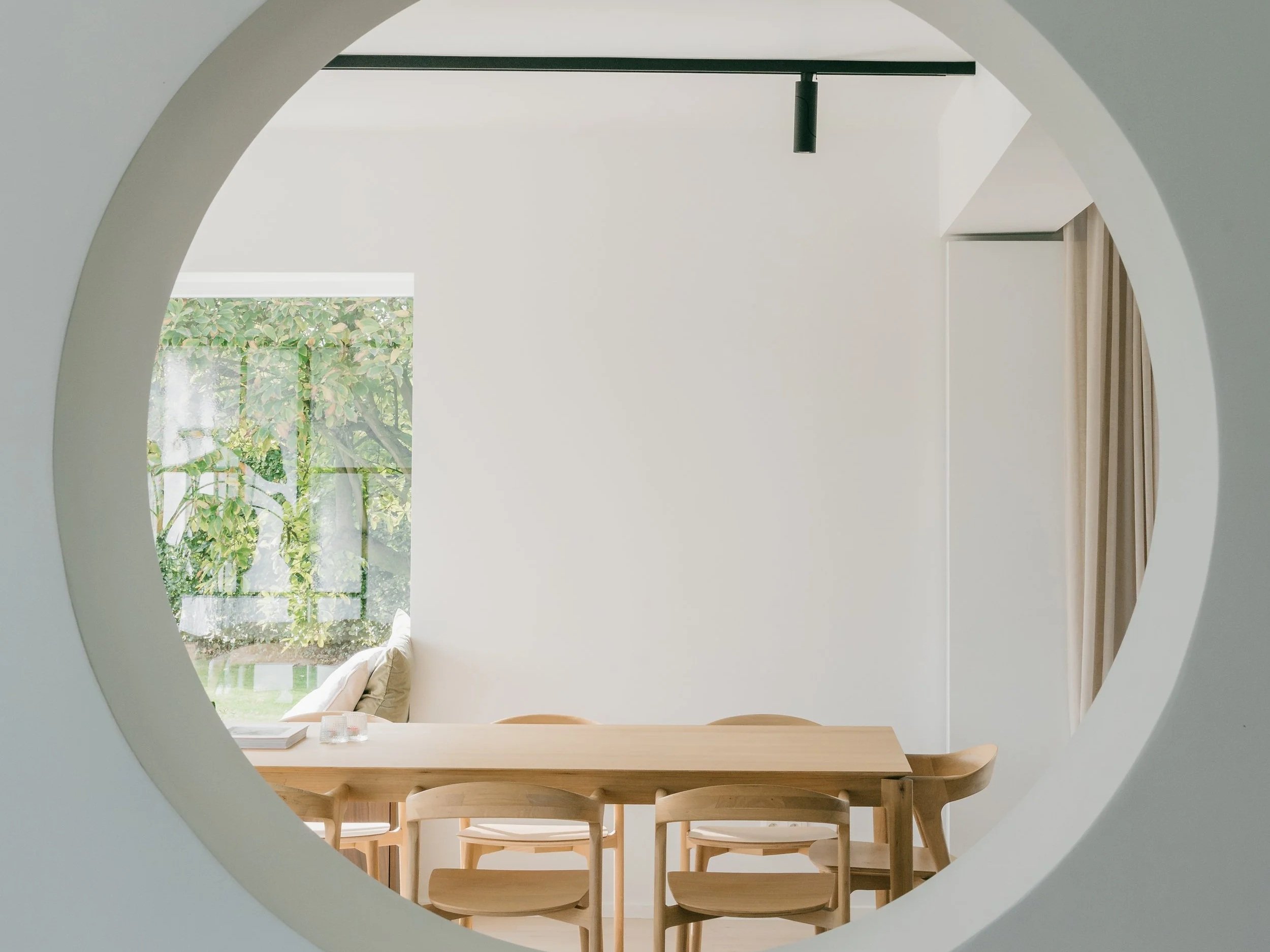 Binnenzicht van een minimalistisch eetkamertafel en stoelen gezien door een ronde opening, met een raam en groene bomen buiten.